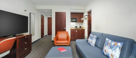 A hotel room with a television, a sofa, an ottoman, a chair, a desk, and a kitchenette.