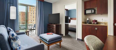 A hotel room with a sofa, ottoman, television, kitchenette, and a balcony with curtains.