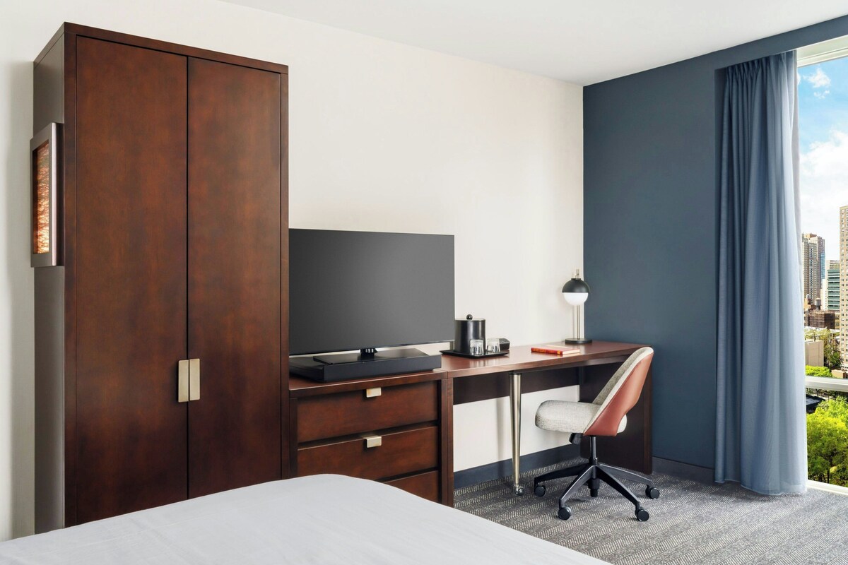 Ein Hotelzimmer mit Bett, Schreibtisch mit Computer, Stuhl, Kleiderschrank und großem Fenster mit Blick auf die Stadt.