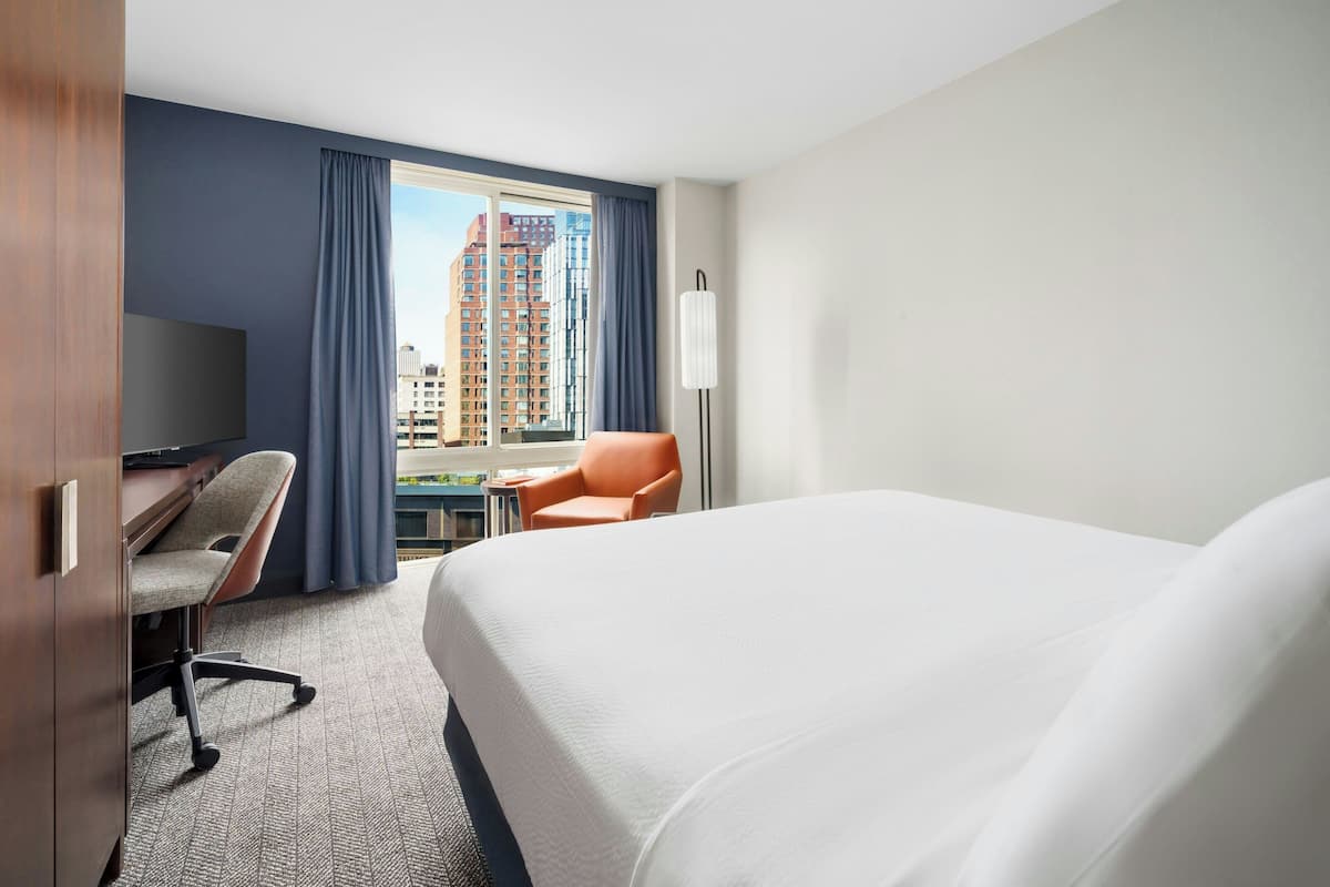 Habitación de hotel con una cama grande, un escritorio con una silla, una mesita y vistas a edificios altos a través del ventanal.