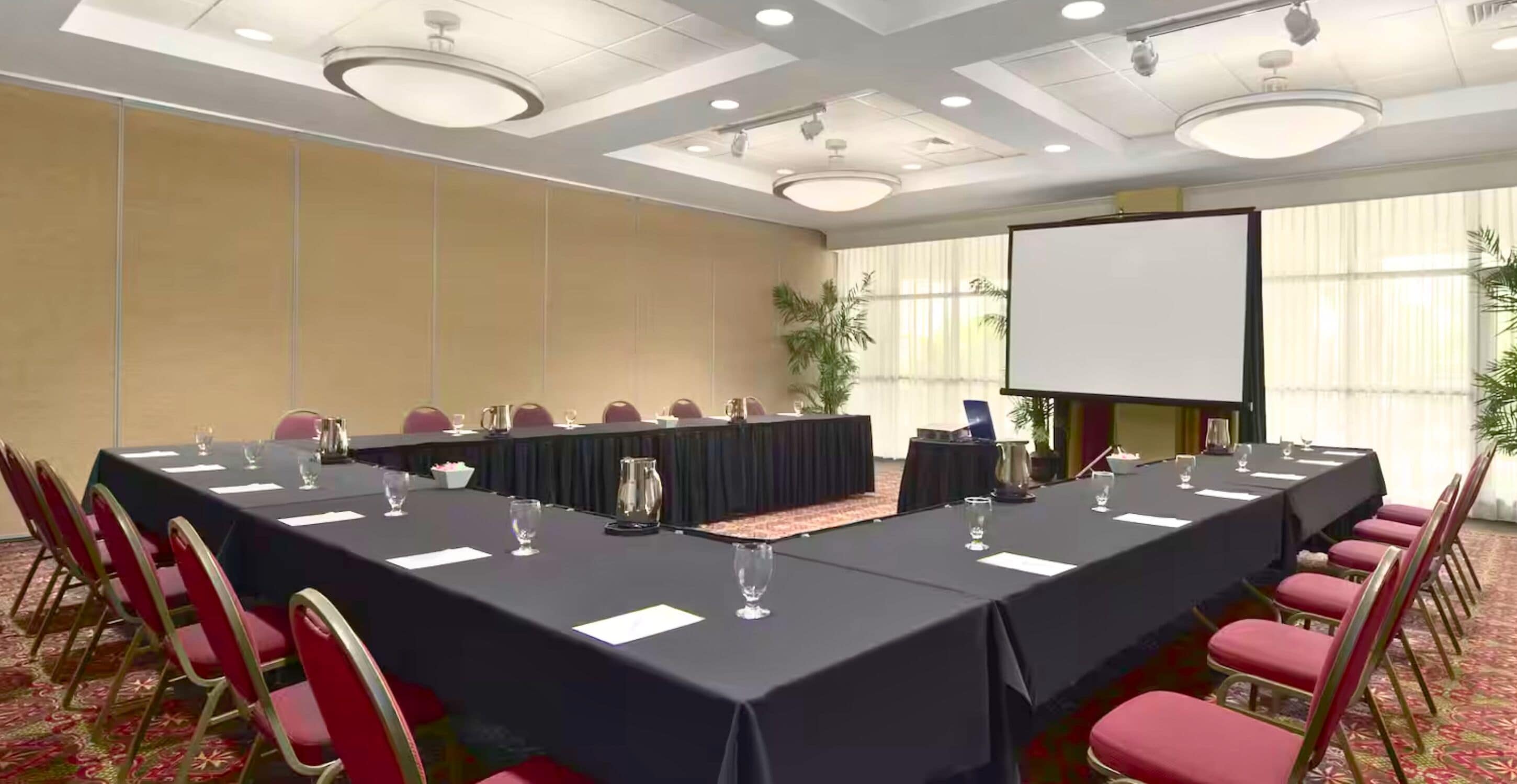 Una sala de conferencias con una larga mesa, sillas rojas, una pantalla de proyector y un amplio ventanal con cortinas.