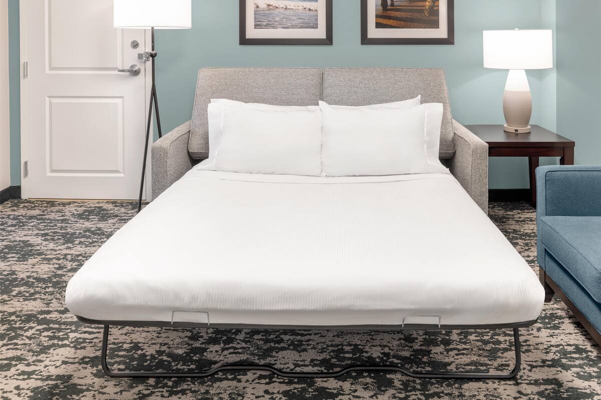A hotel room with a bed, a chair, a nightstand, two lamps, and framed pictures on the wall.