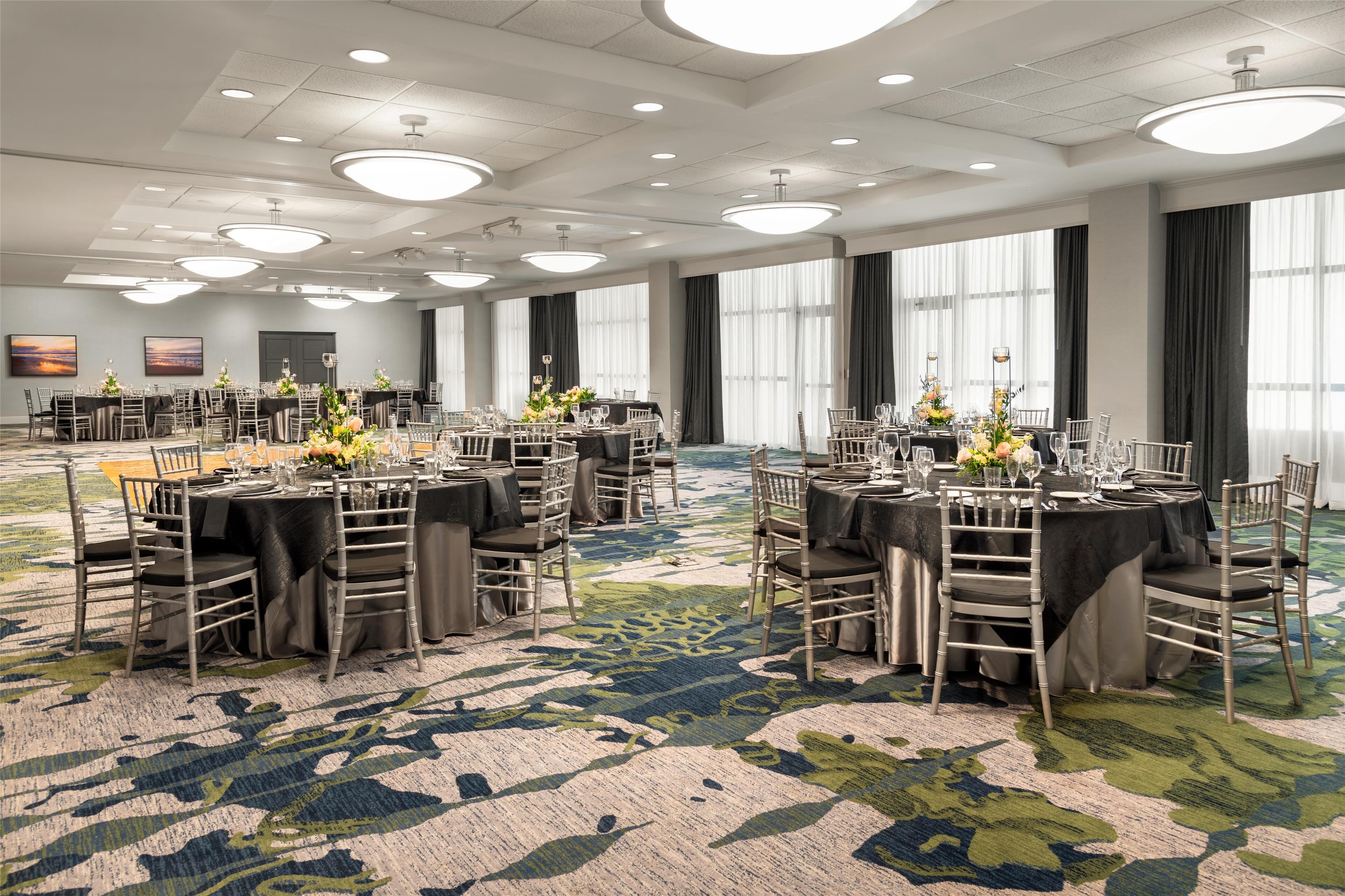 A large conference room with multiple round tables, each set with chairs and centerpieces. The room has a carpeted floor and large windows with curtains.