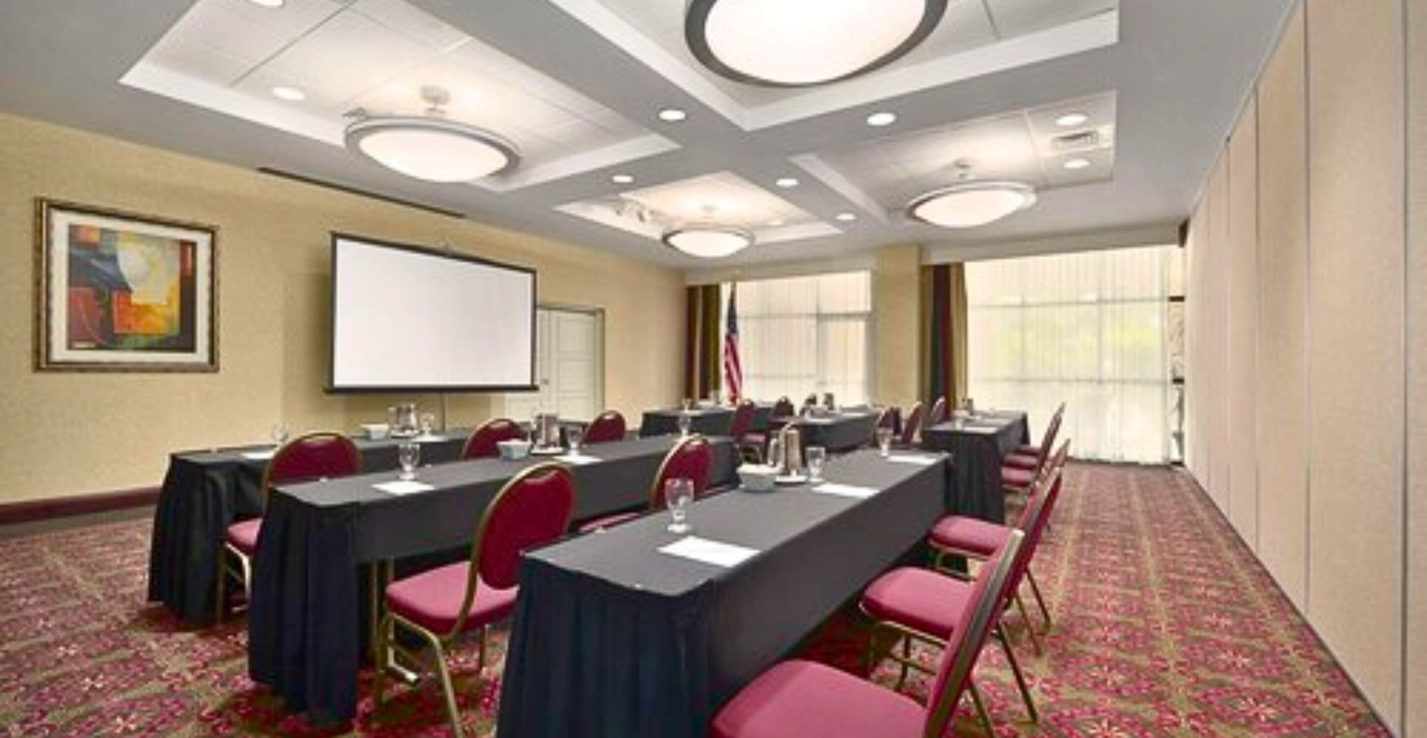 A conference room with long tables, red chairs, a projector screen, and a carpeted floor.