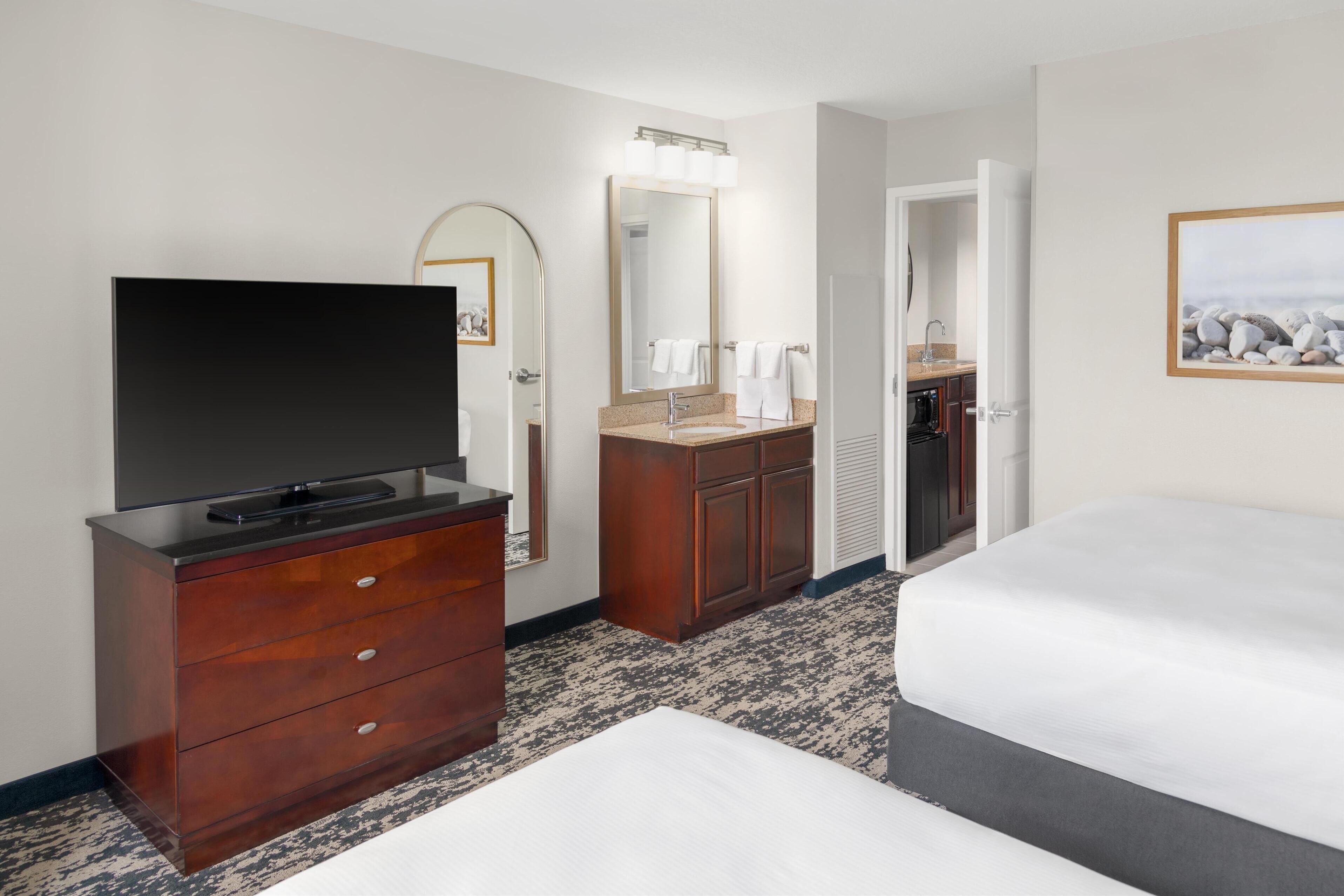 Une chambre d’hôtel avec un téléviseur à écran plat posé sur une commode en bois, un grand miroir au-dessus de la coiffeuse et un lit avec un matelas gris.