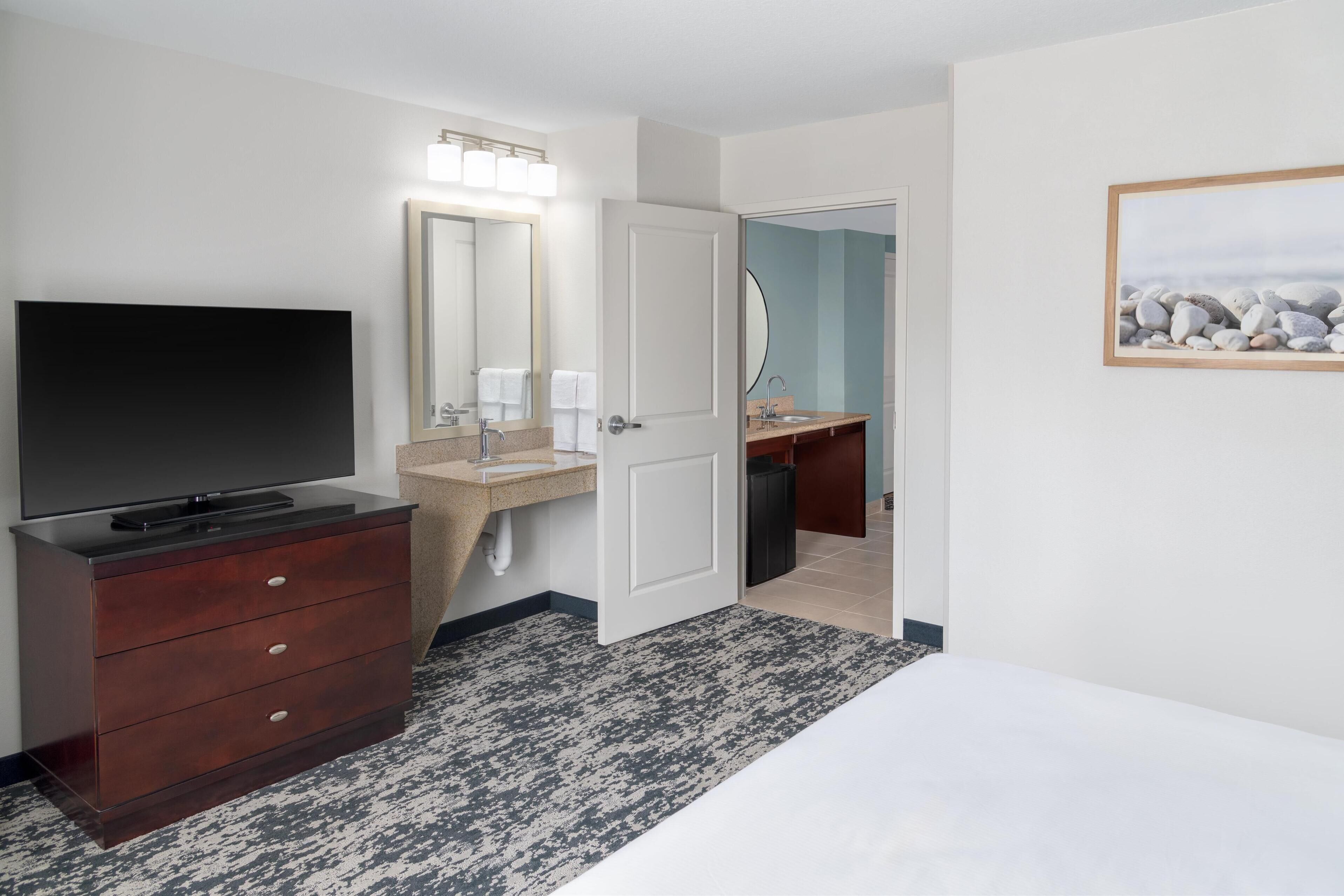 Habitación de hotel con televisor sobre un tocador de madera, baño con lavamanos y espejo, y un cuadro en la pared.