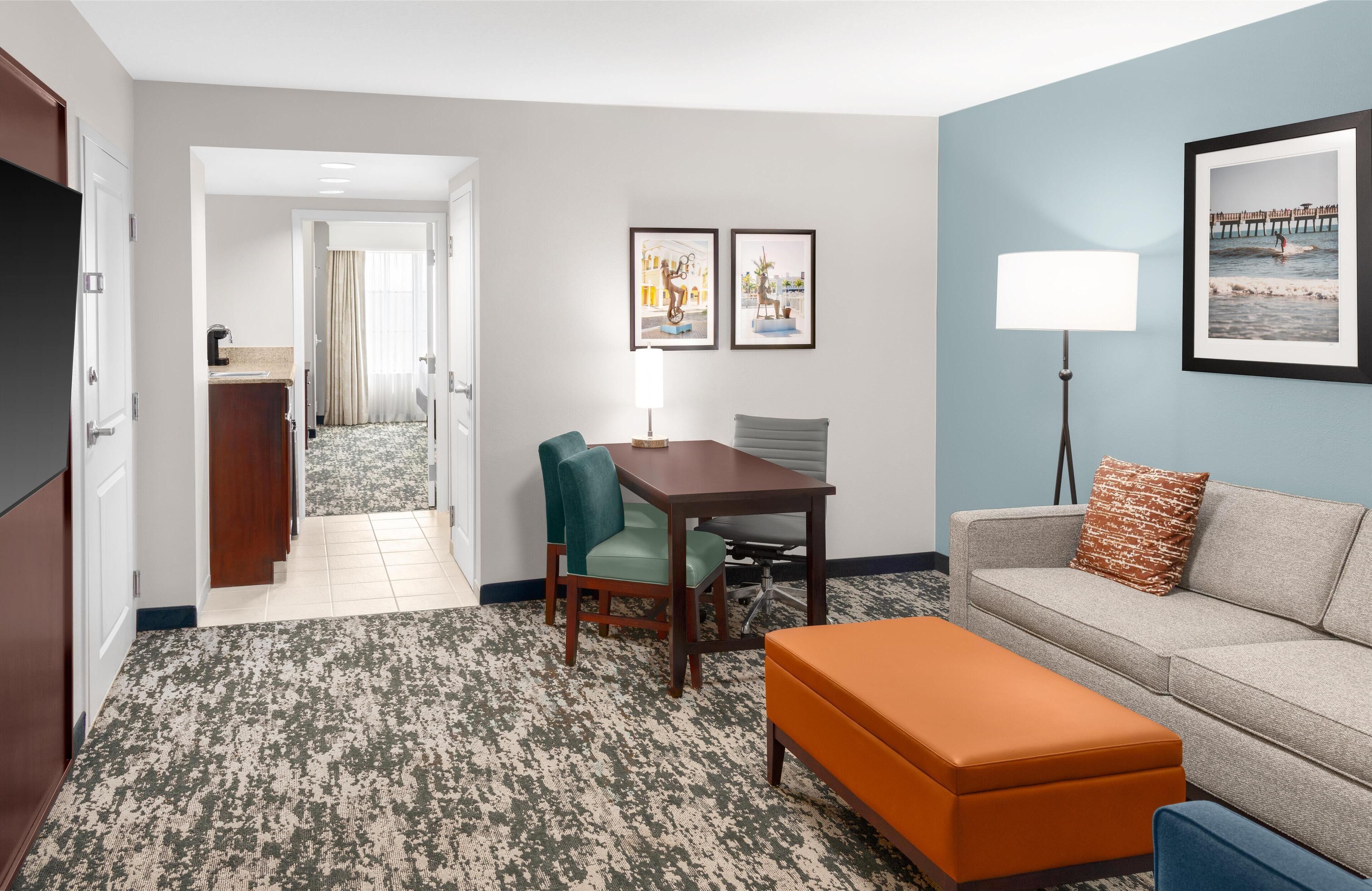 Habitación de hotel con zona de comedor, sofá, reposapiés, lámpara y obras de arte en las paredes.