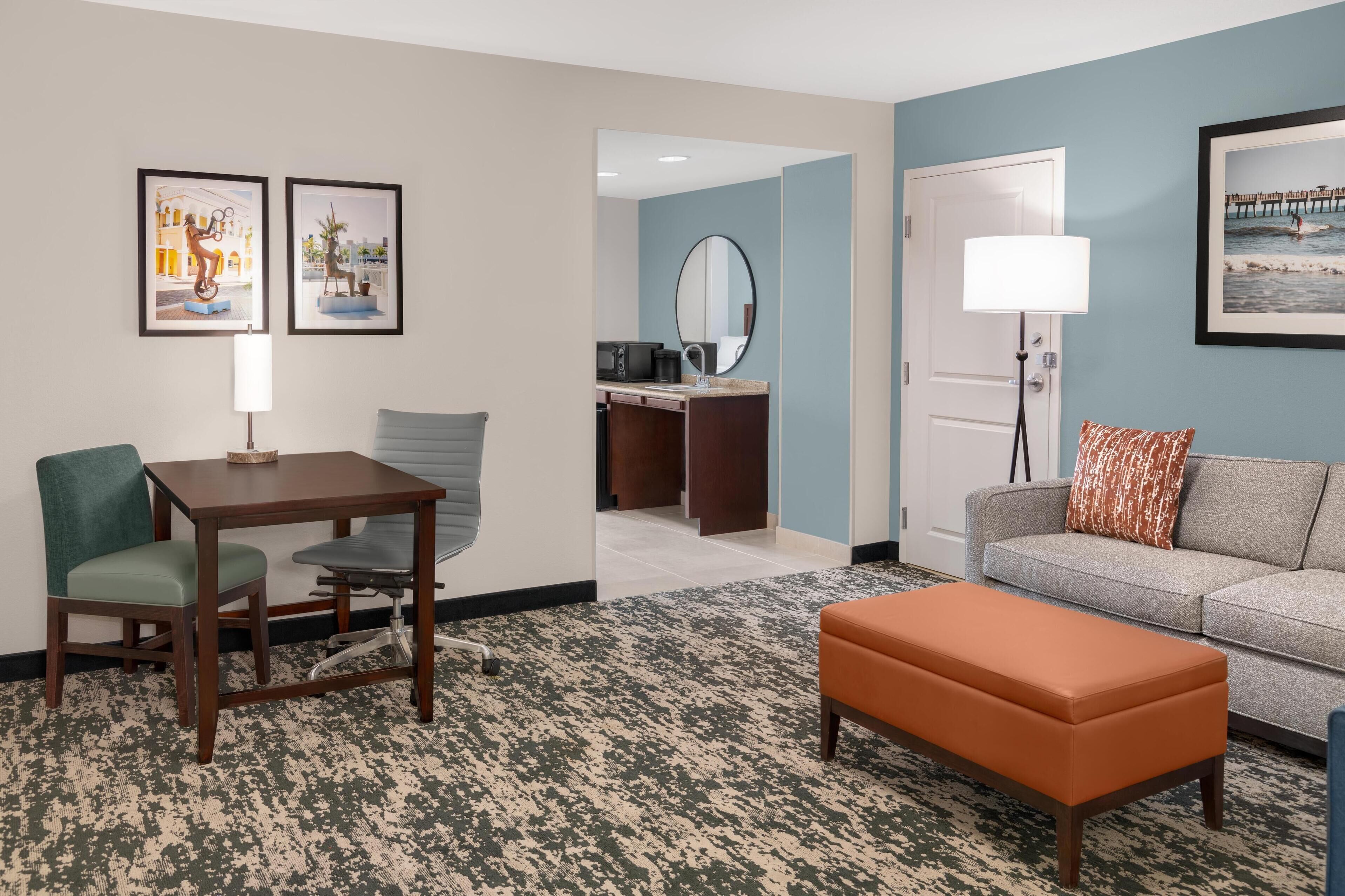 A hotel room with a desk, chair, sofa, and ottoman. There is a mirror, a lamp, and artwork on the walls.