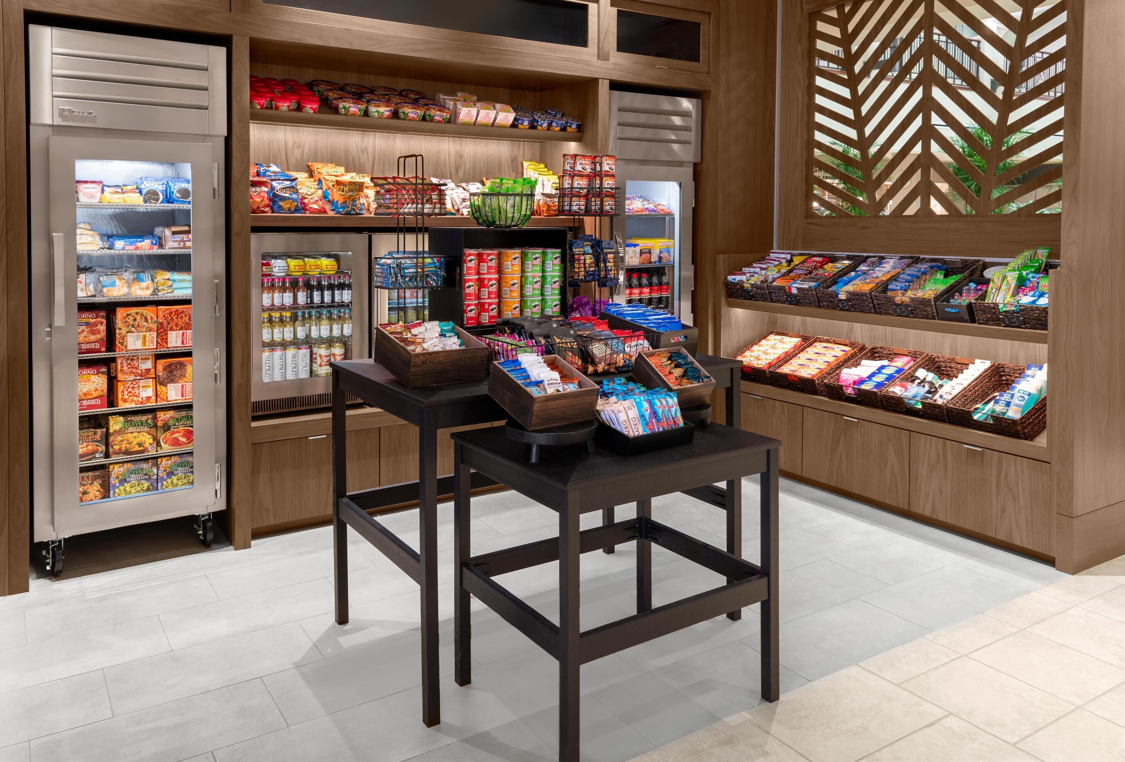 A grocery store with refrigerated sections, shelves stocked with snacks, and a display table with packaged goods.