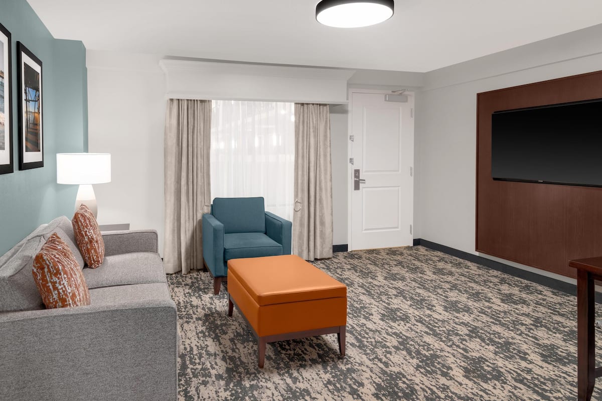 A hotel room with a grey sofa, an orange ottoman, a blue armchair, a flat-screen TV, and a large window with curtains.