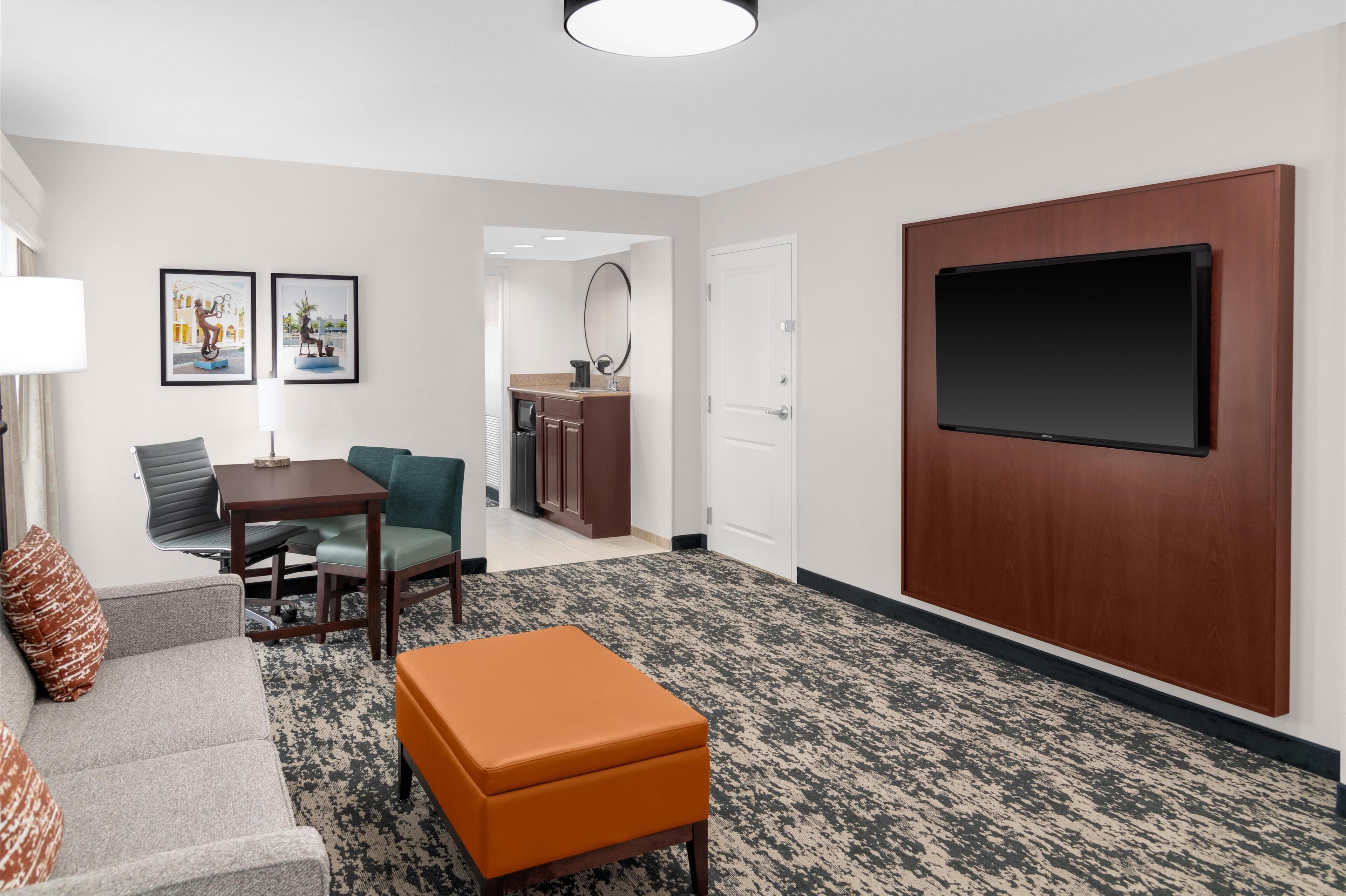A hotel room with a gray sofa, an orange ottoman, a dining table with chairs, a kitchen area, and a large flat-screen TV mounted on a wooden panel.