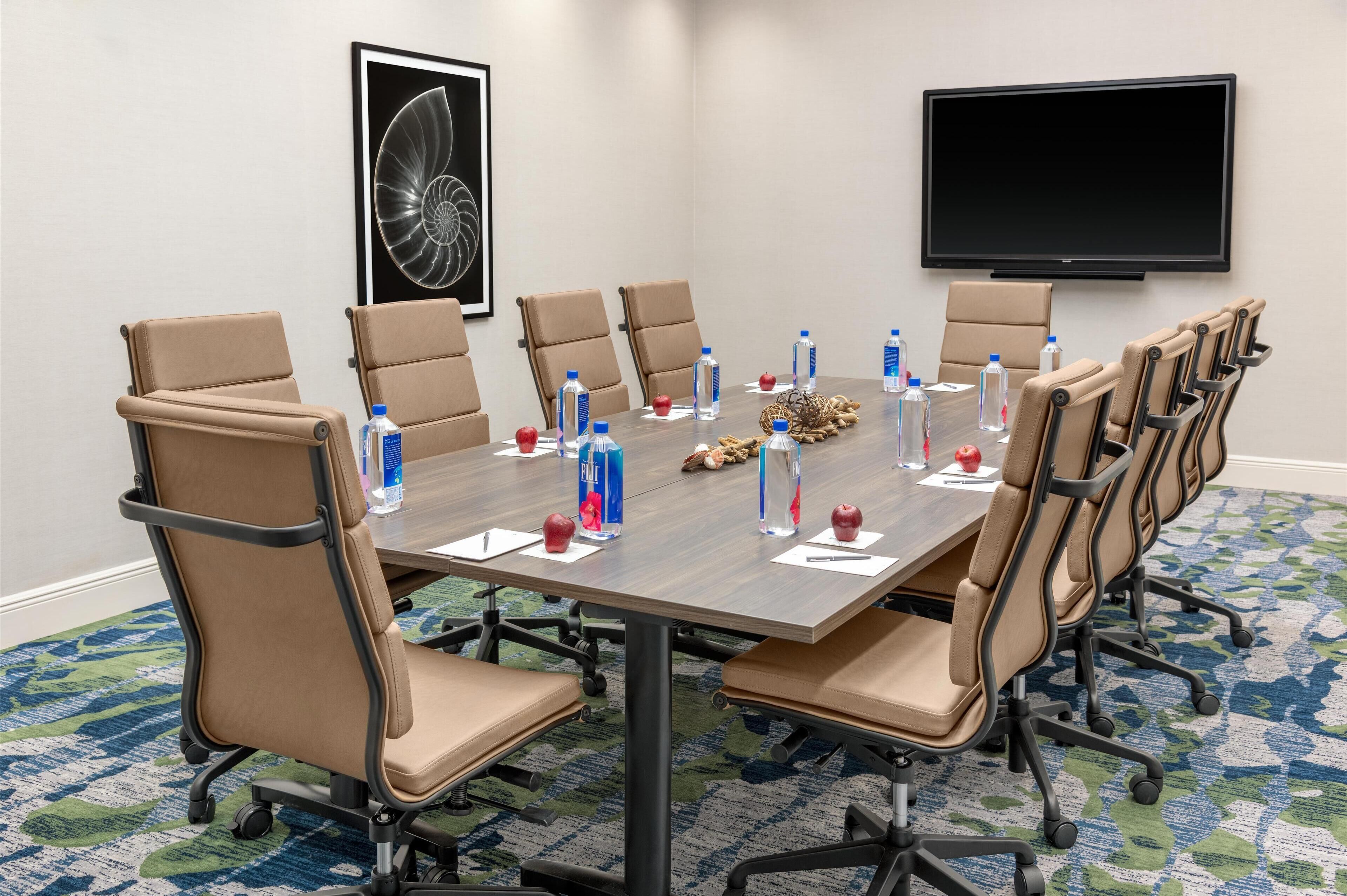 A conference room with a long table, beige chairs, water bottles, and a flat-screen TV on the wall.