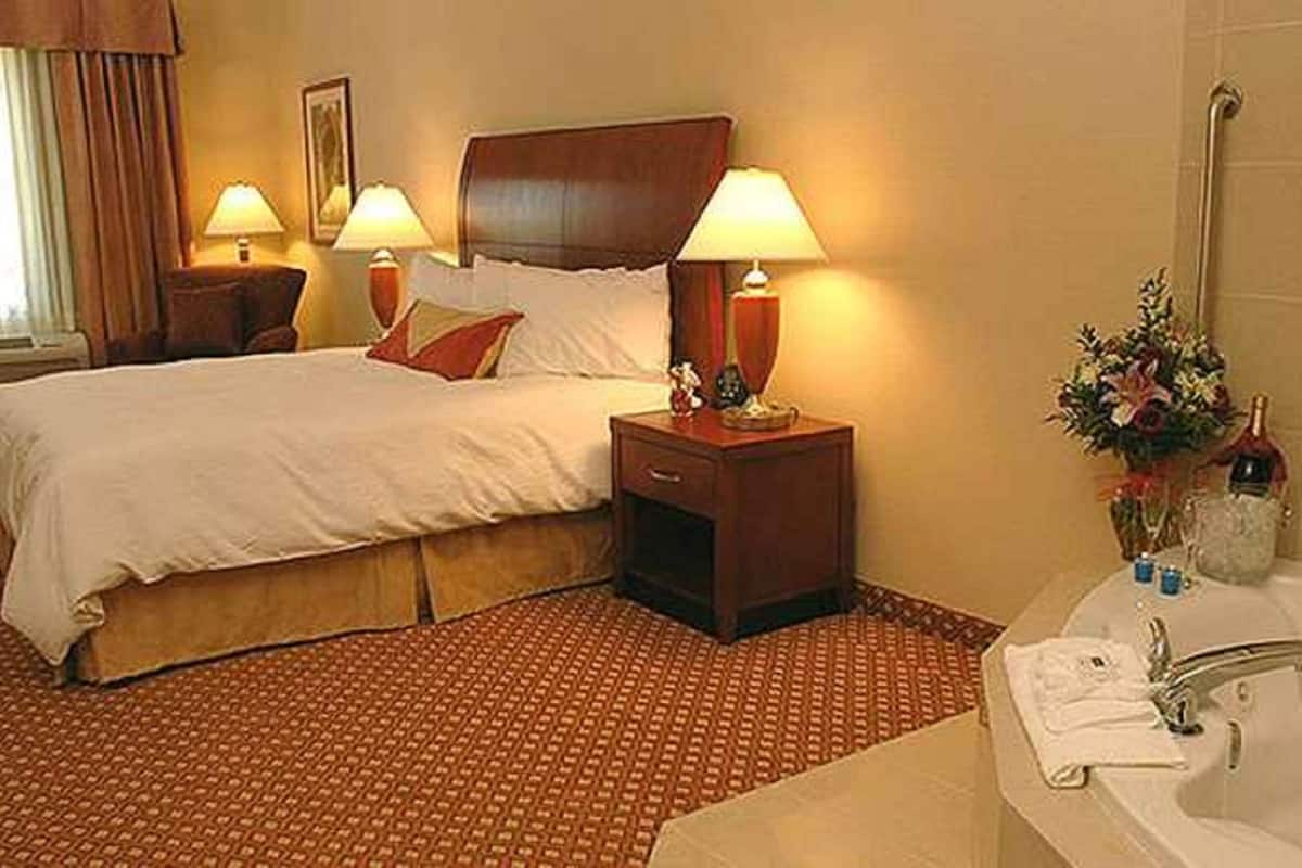 Une chambre d’hôtel avec un grand lit, deux tables de chevet avec des lampes, une salle de bain avec une baignoire et un vase de fleurs.