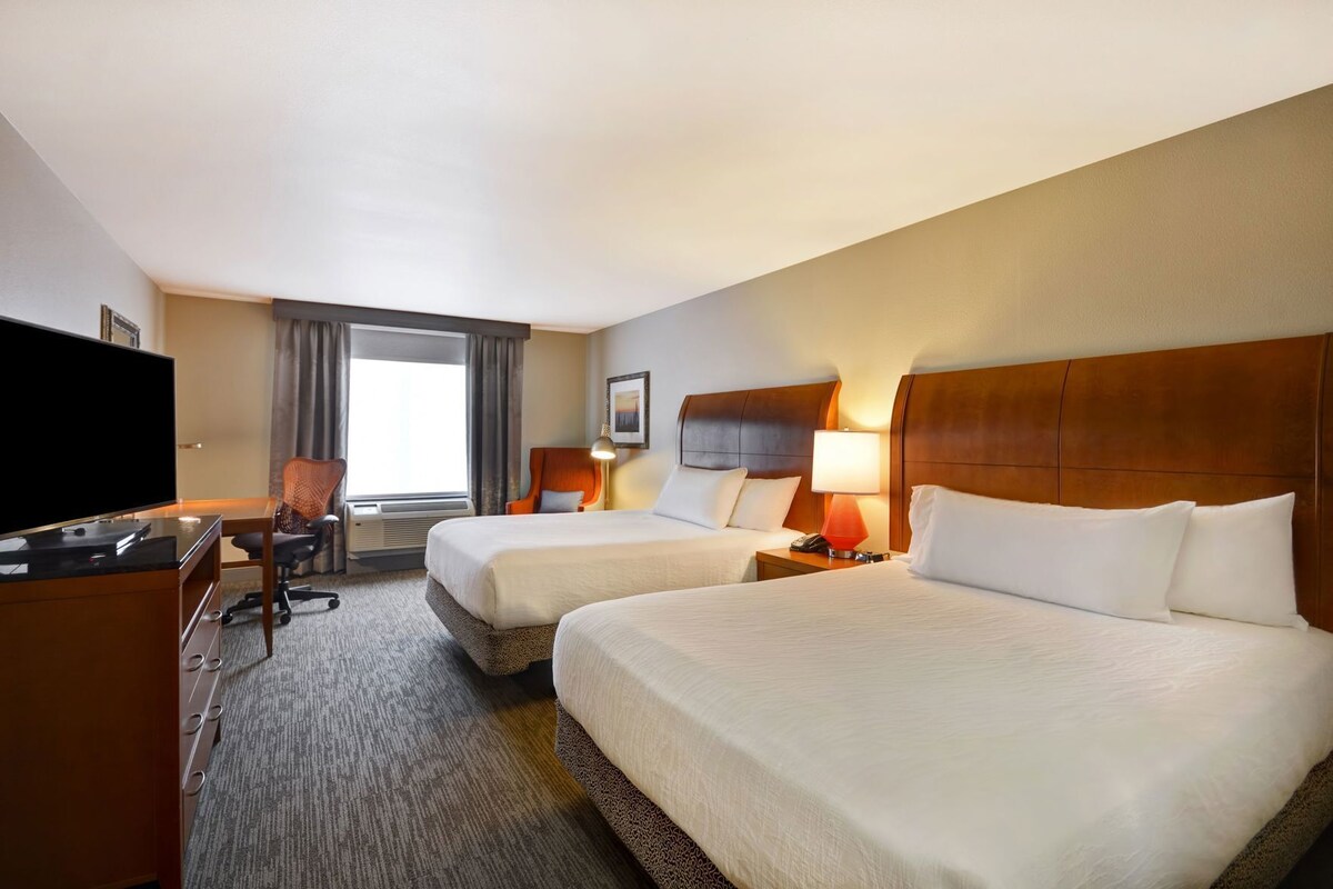 A hotel room with two beds, a desk with a TV, a chair, and a window with curtains.