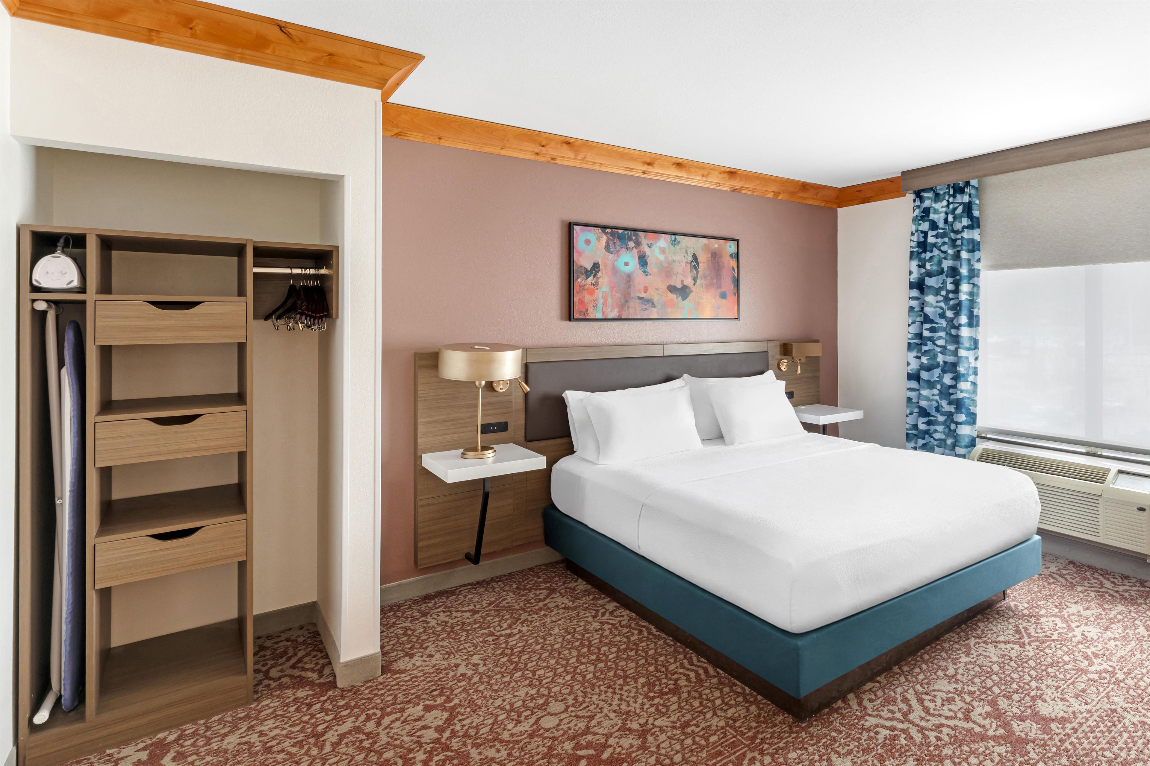 A hotel room with a bed, a wardrobe, a nightstand, a lamp, a painting, and a window with curtains.
