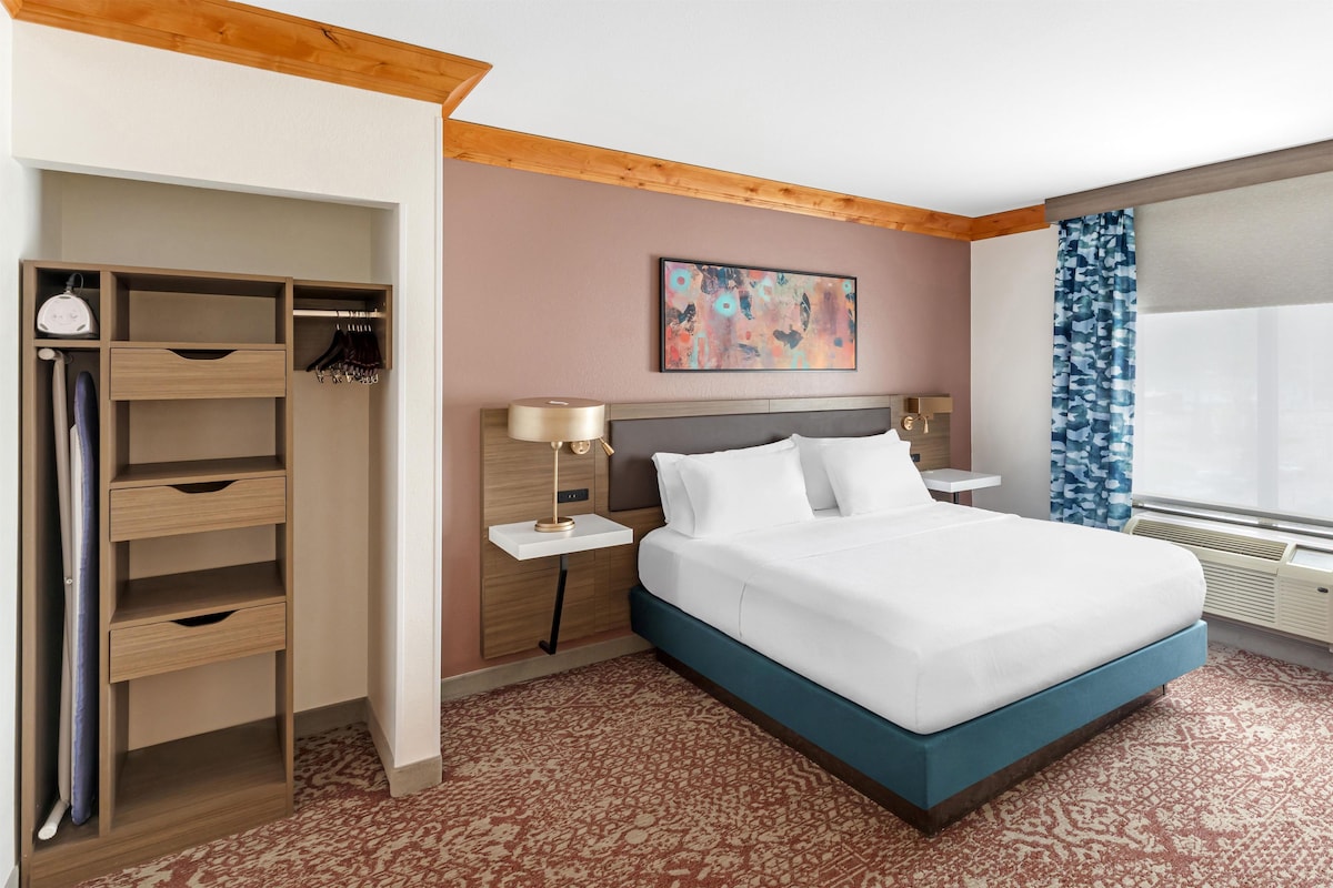 Habitación de hotel con cama, armario, mesita de noche, lámpara, un cuadro y ventana con cortinas.