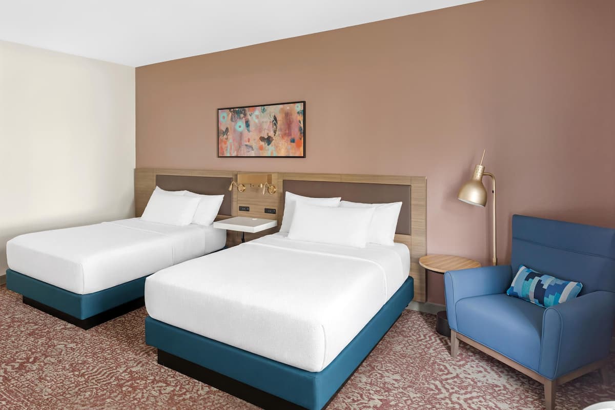 A hotel room with two beds, a bedside table, a lamp, and a blue armchair.