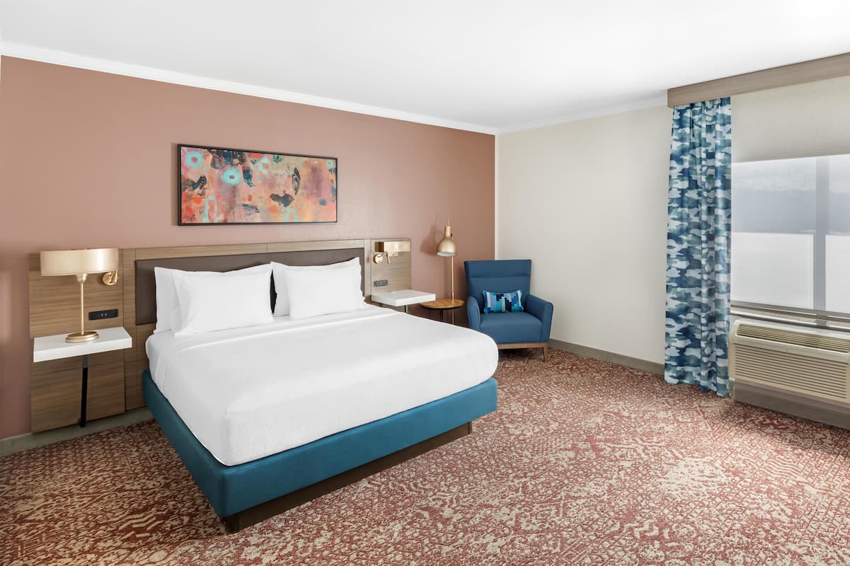 A hotel room with a bed, a chair, a nightstand, a lamp, a painting, and a window with curtains.