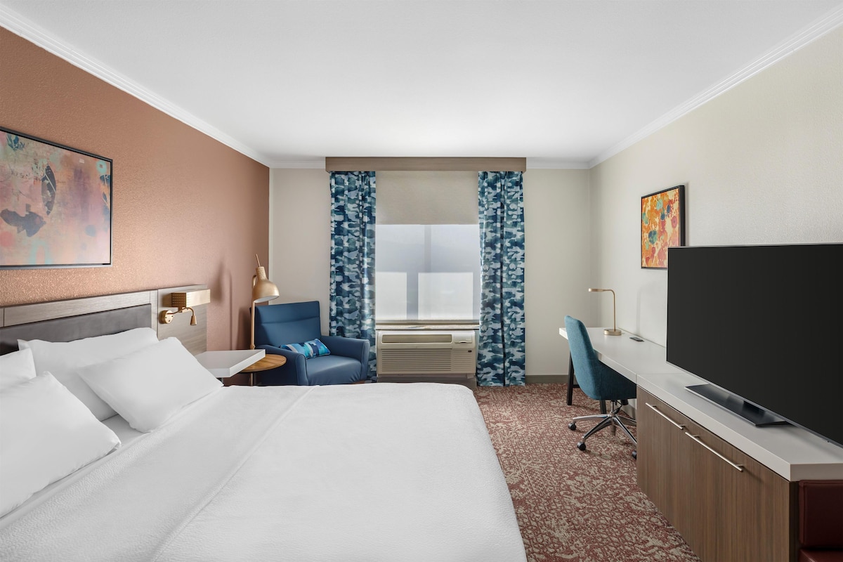 Habitación de hotel con cama, escritorio, silla, televisor y ventana con cortinas.