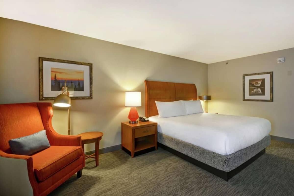 A hotel room with a bed, a chair, a nightstand, a lamp, and two framed pictures on the walls.