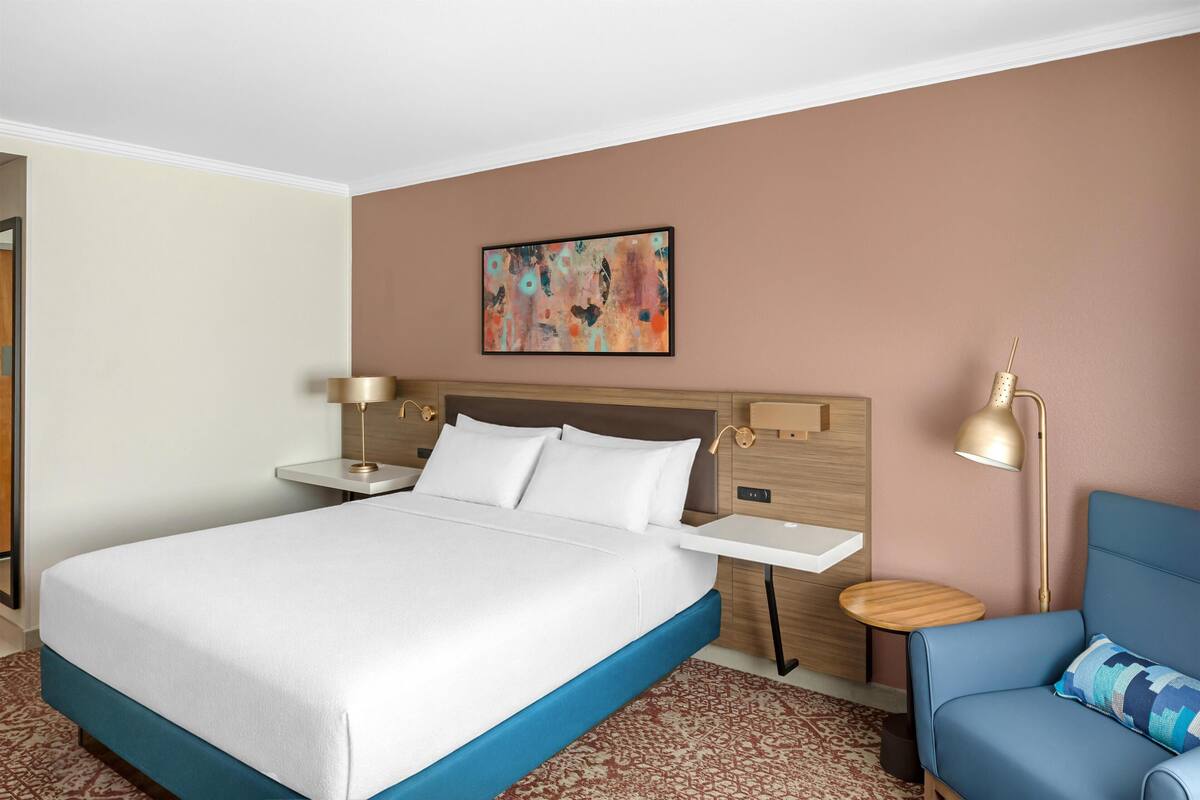 A hotel room with a bed, bedside tables, a lamp, a chair, and a painting on the wall.
