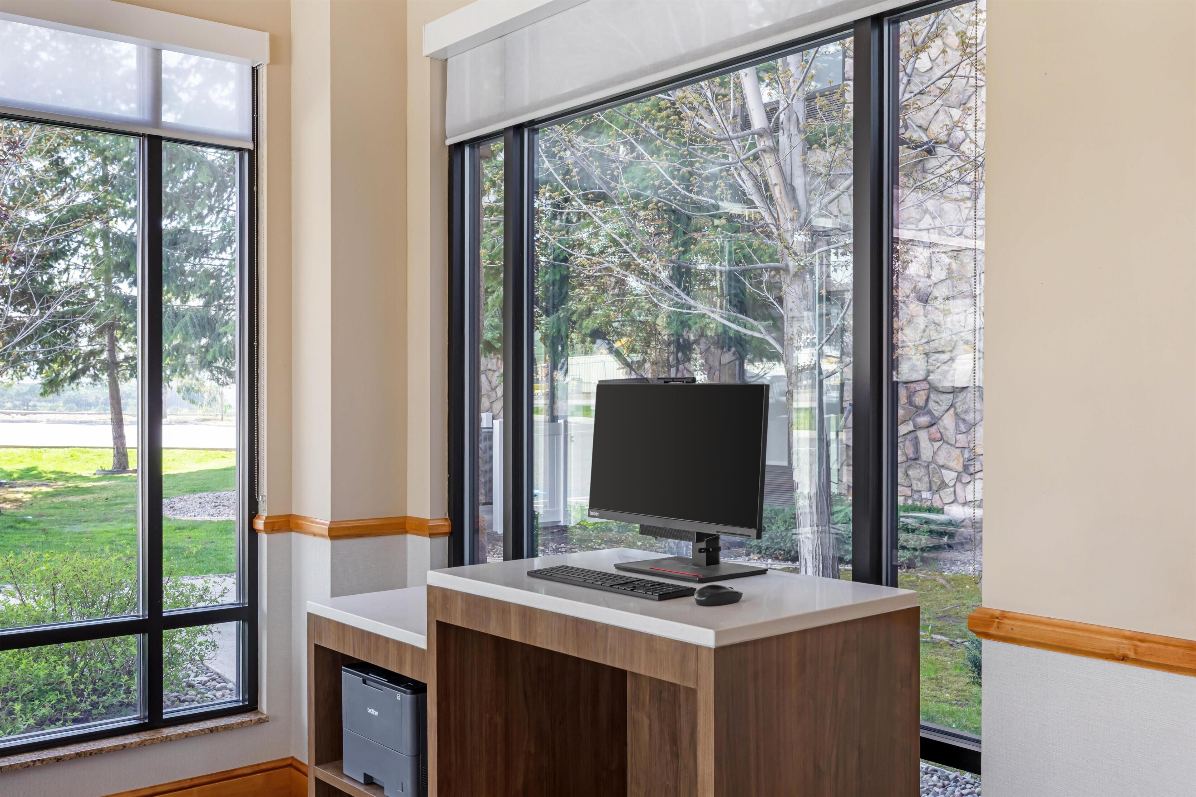 A modern office desk with a computer, positioned near large windows with a view of trees and a stone wall.