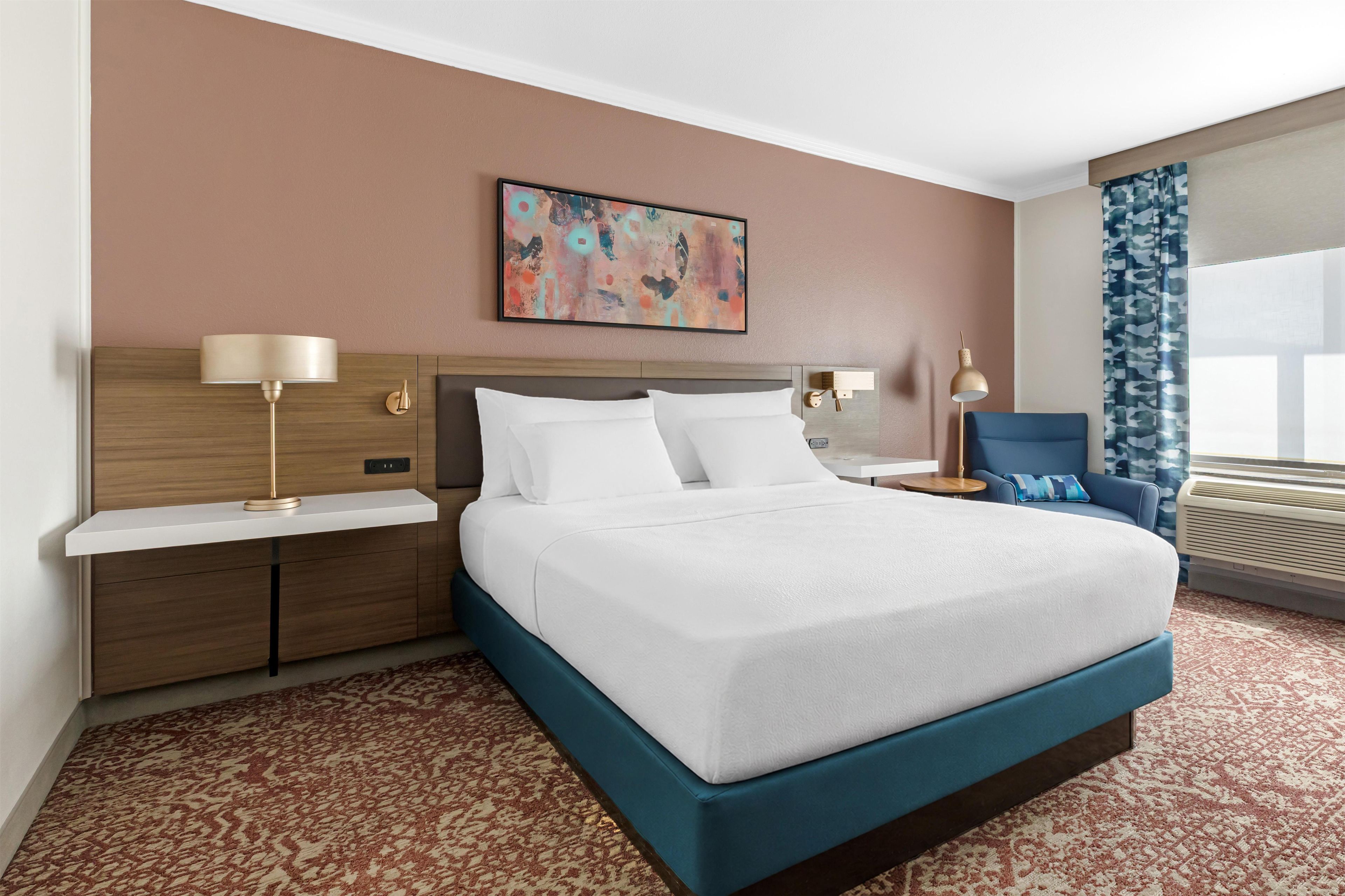 Habitación de hotel con cama, mesitas de noche, lámpara, un cuadro, una silla y una ventana con cortinas.