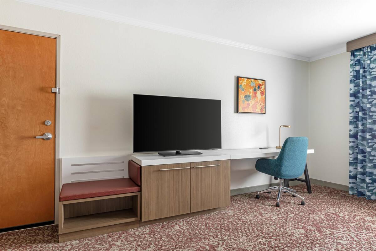 A hotel room with a wooden desk, a television, a chair, a bench, a painting, and a patterned carpet.