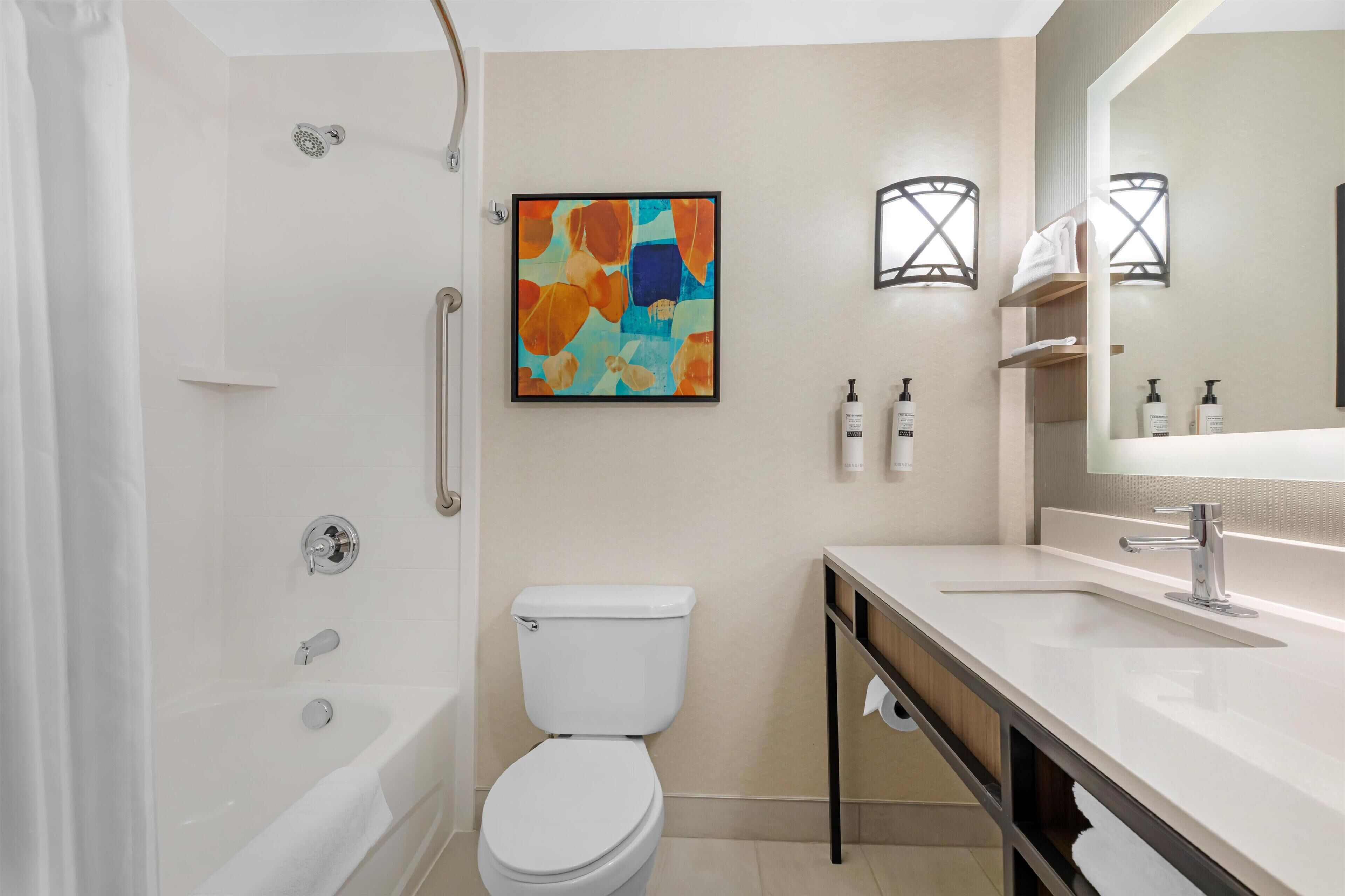 A modern bathroom with a bathtub, toilet, and sink, featuring a large mirror and abstract art on the wall.