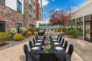 A long dining table set outdoors with chairs, surrounded by a stone building and a glass structure. - Hilton Garden Inn Missoula (Missoula)