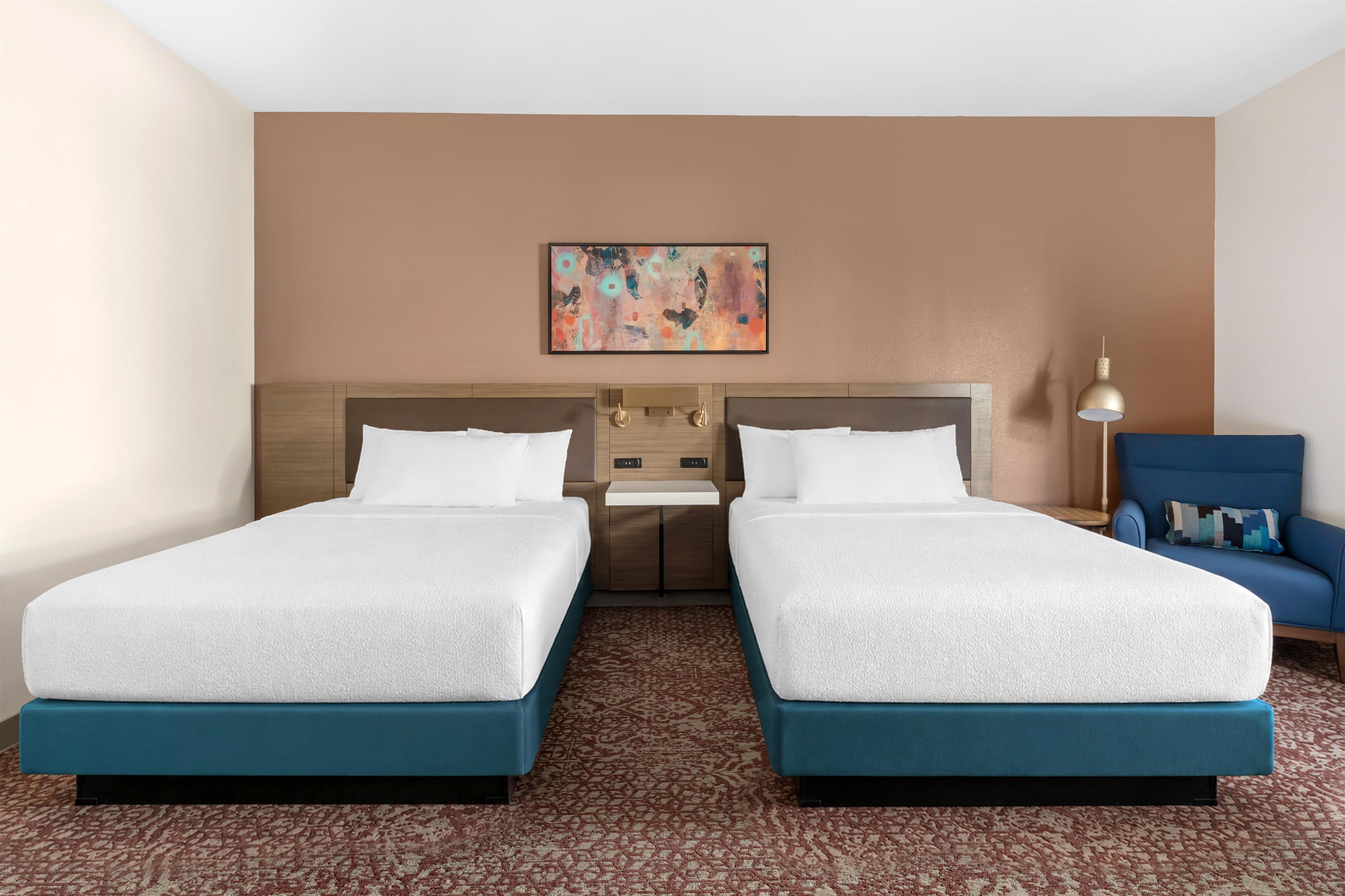 A hotel room with two beds, a headboard, a nightstand, a lamp, and a blue chair.