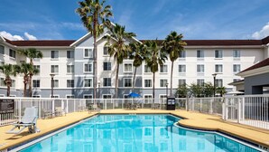 Una piscina con valla blanca, palmeras y un edificio de hotel al fondo.