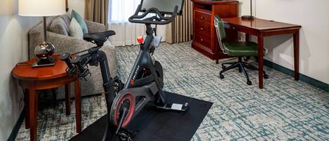 A hotel room with a stationary bike, a desk, a television, and a couch.