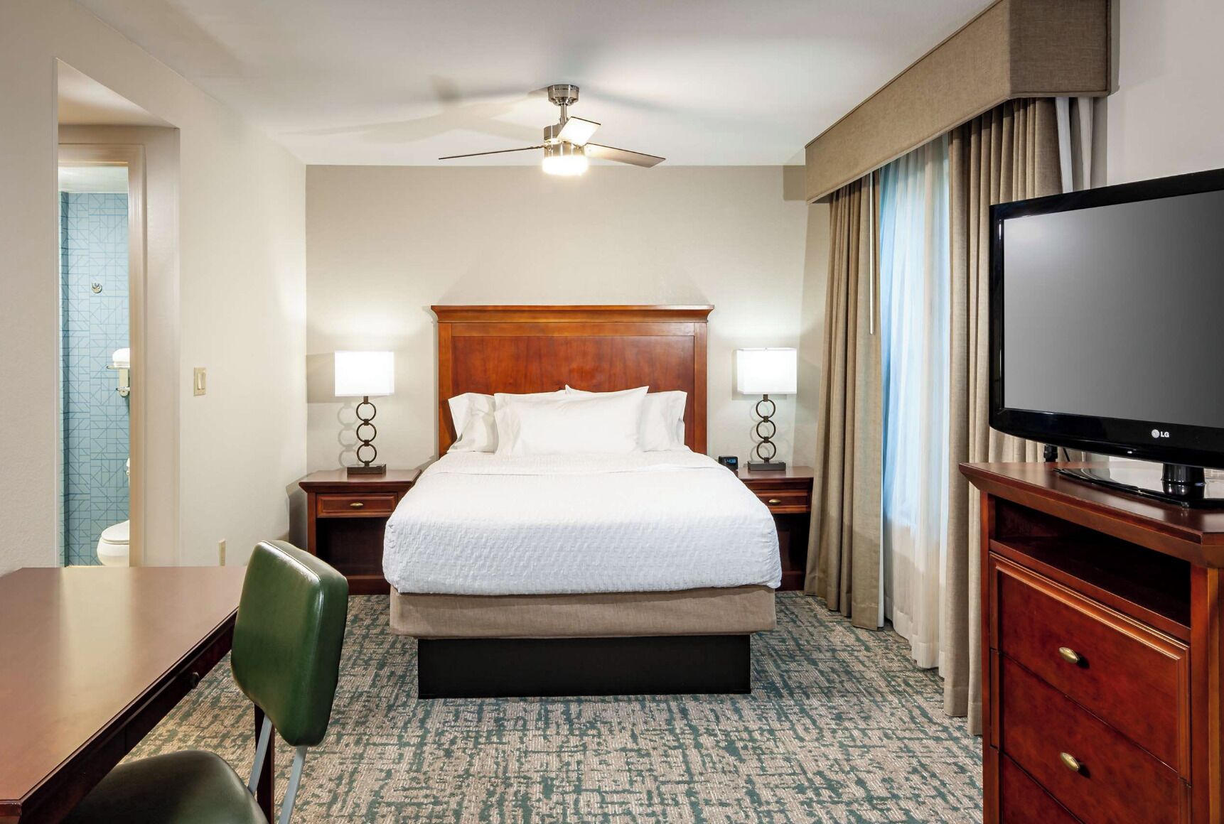 Une chambre d’hôtel comprenant un lit, deux tables de chevet avec des lampes, une télévision, un bureau avec une chaise et un sol recouvert de moquette.