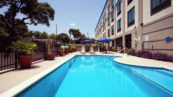 Una piscina con superficie d'acqua cristallina e blu, circondata da piante in vaso, e un edificio con molte finestre e un tendone blu.
