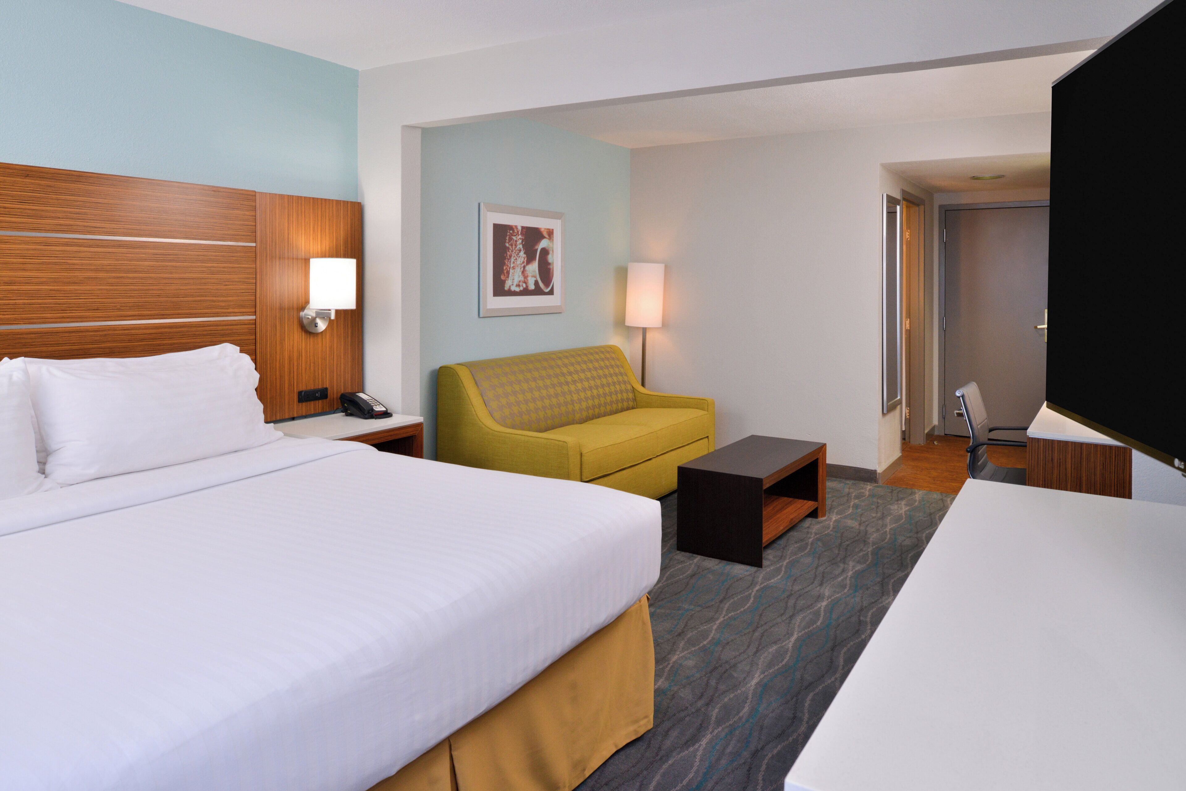 Habitación de hotel con una cama grande, un sofá, una mesita, un escritorio, un televisor y una puerta.