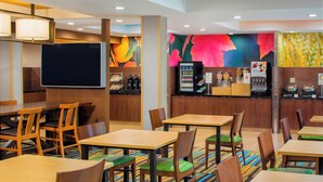 Rebedor d'hotel amb taules i cadires de fusta, un televisor de pantalla plana gran, una màquina de cafè i un dispensador de suc de fruita.
