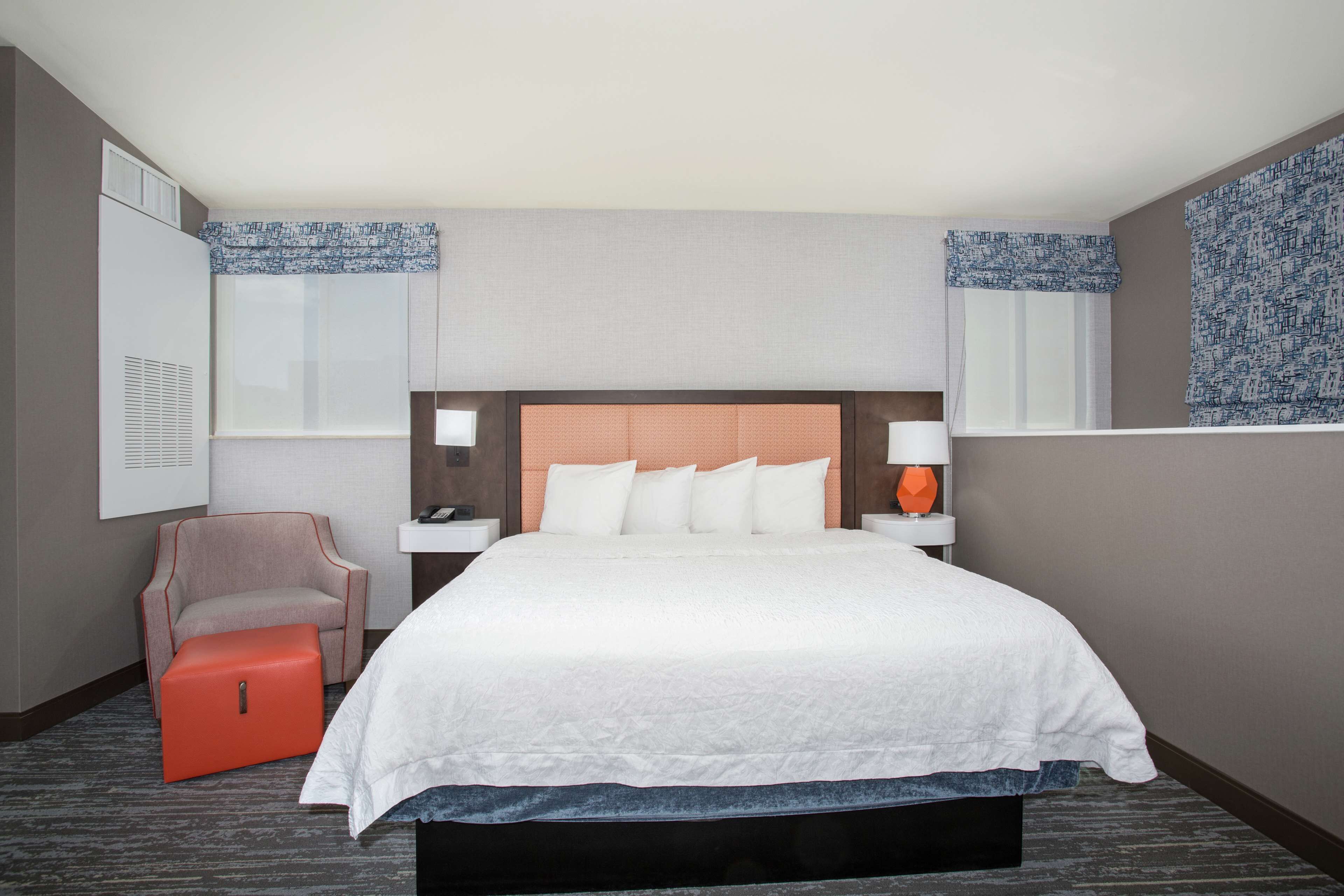 A hotel room with a large bed, a chair, a nightstand, and a window with patterned curtains.