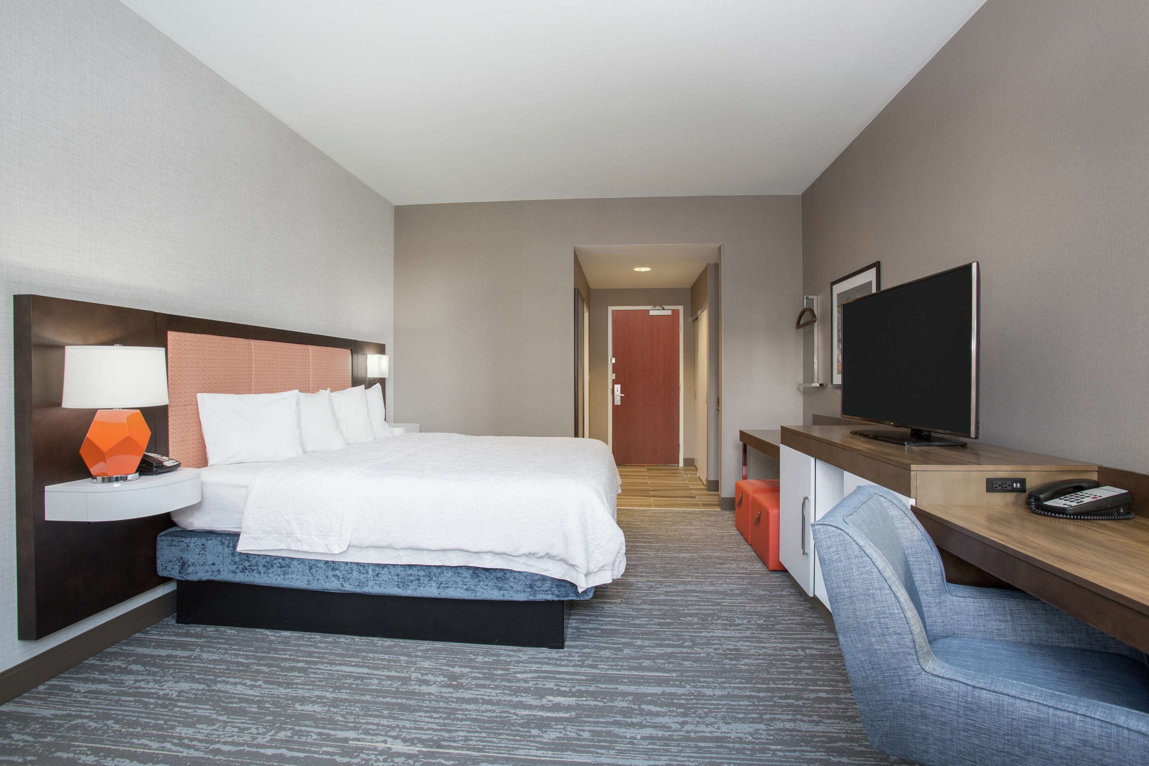 A hotel room with a bed, a desk with a television, a chair, and a nightstand with a lamp.