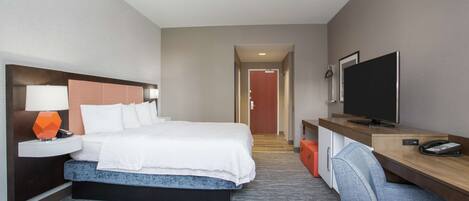 A hotel room with a bed, a desk with a television, a chair, and a nightstand with a lamp.