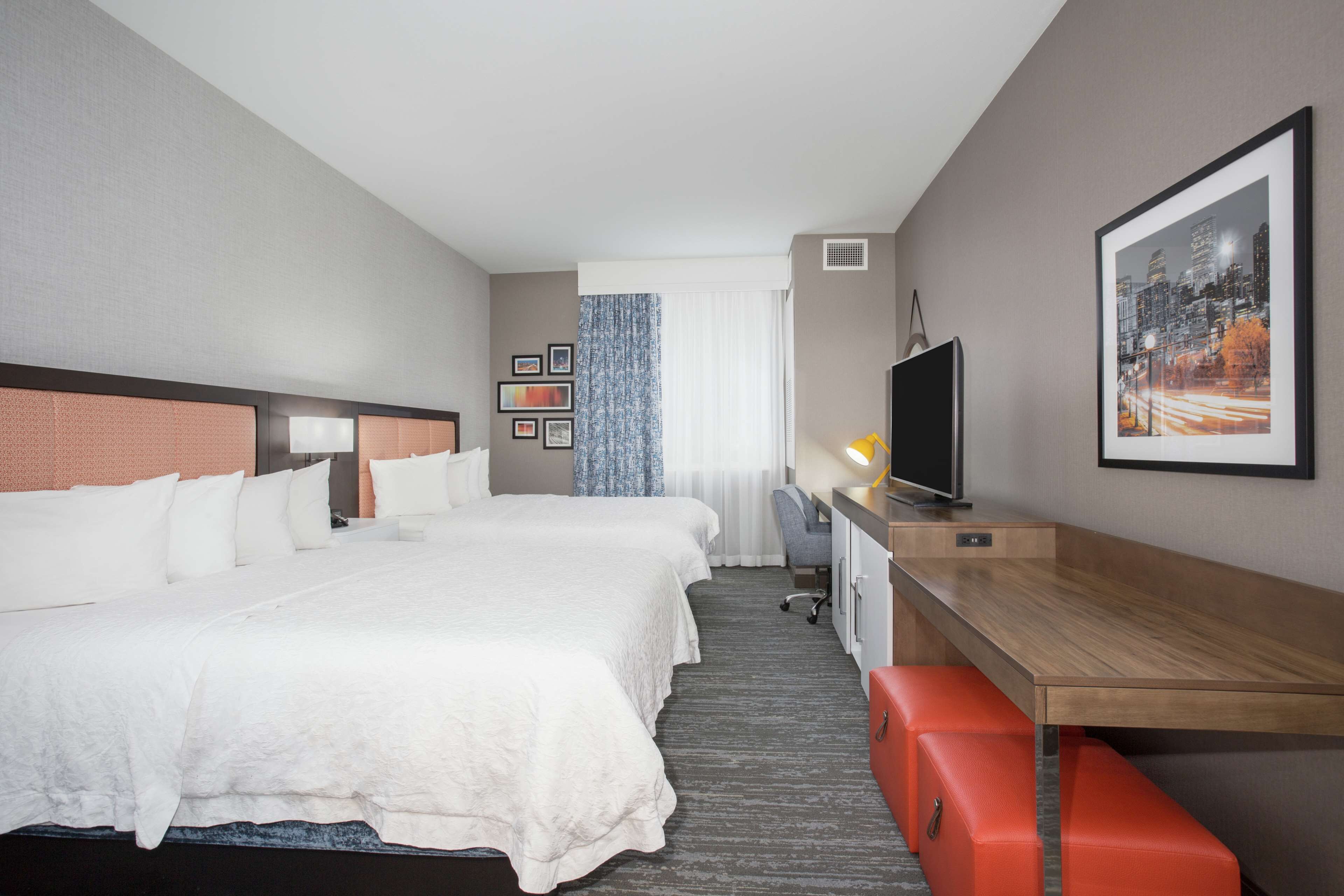 A hotel room with two beds, a desk, a television, and a cityscape painting.