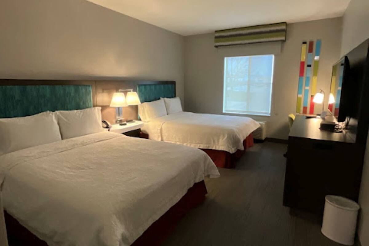 A hotel room with two beds, a desk, a TV, and a window.