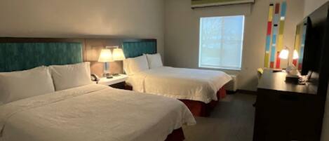 A hotel room with two beds, a desk, a TV, and a window.