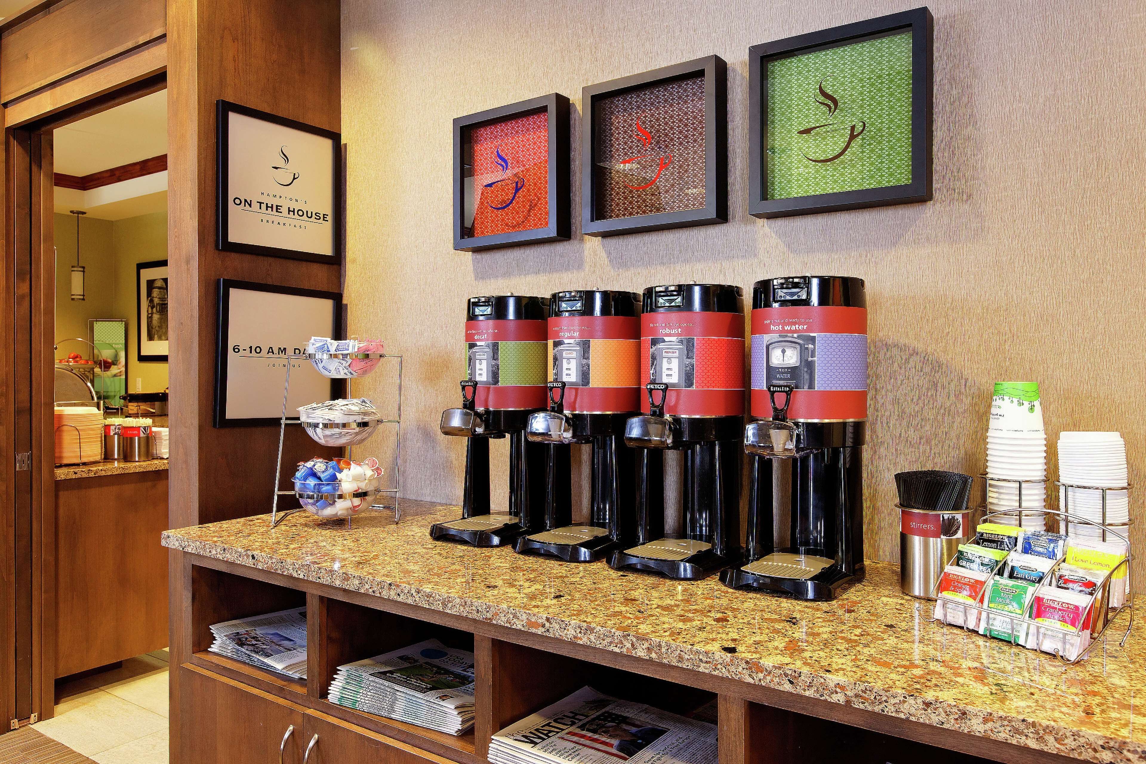 A coffee station with multiple coffee makers, a newspaper rack, and framed artwork on the wall.