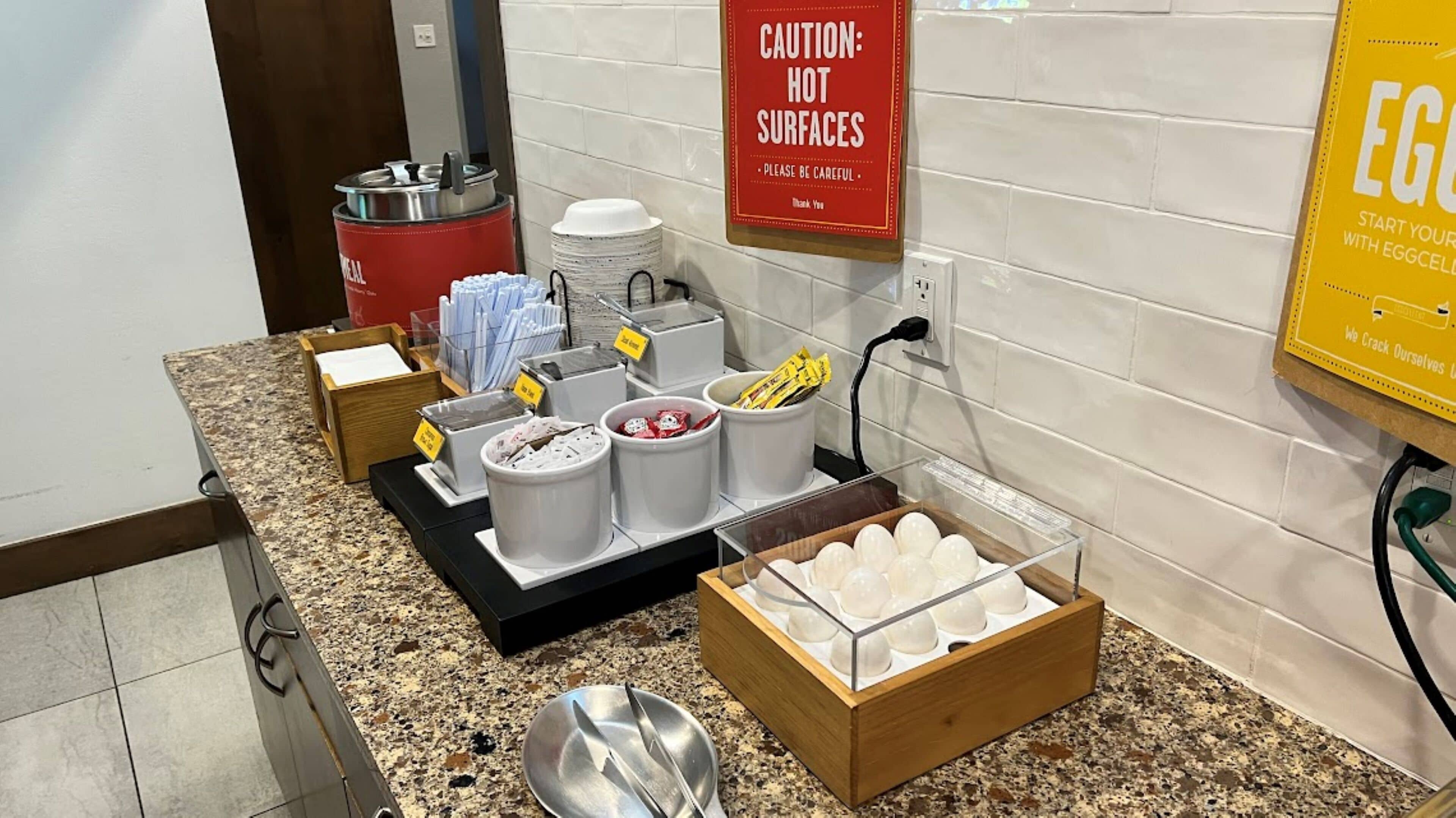 A breakfast station with various food items and utensils, a caution sign for hot surfaces, and a promotional sign for eggs.