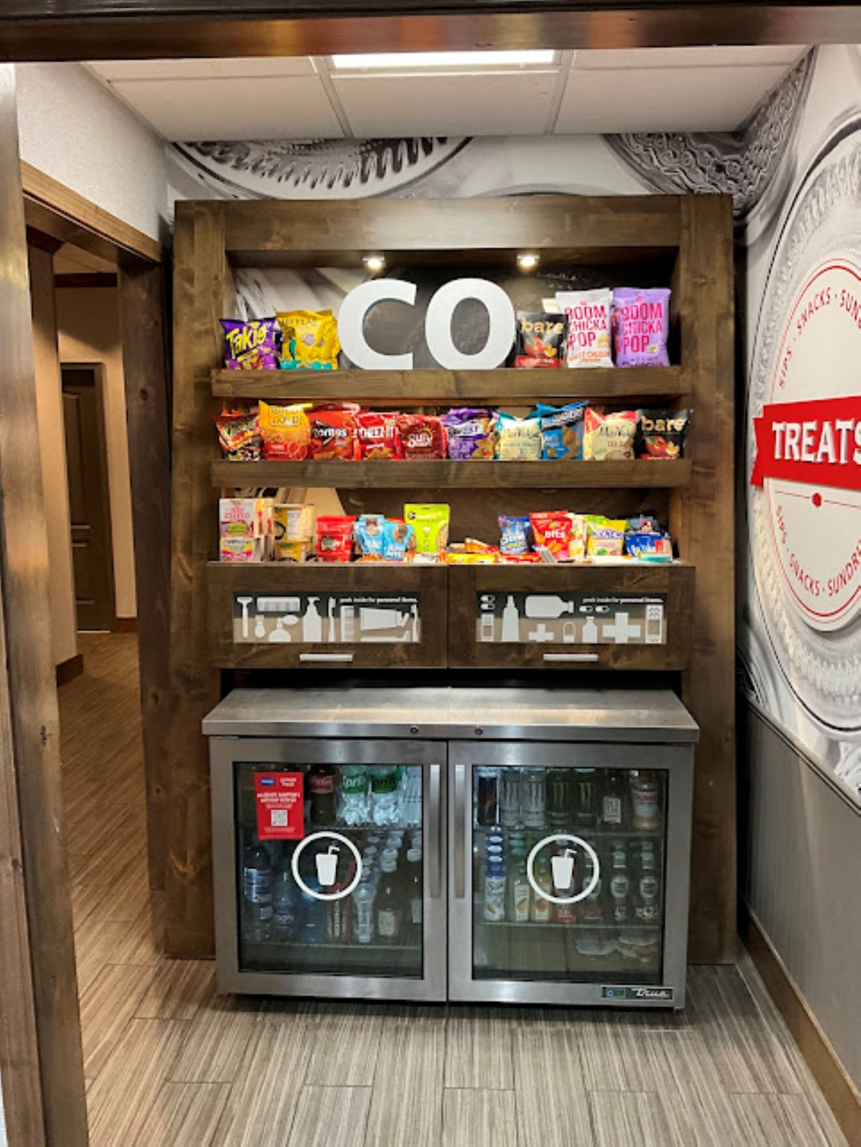 A snack display with a 'CO' sign above, shelves of colorful snack bags, and two refrigerated beverage dispensers.