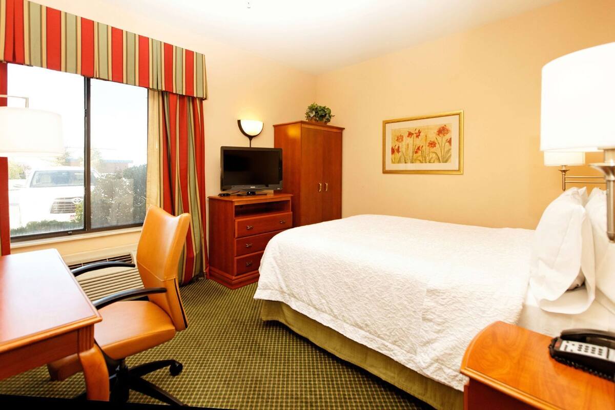 A hotel room with a bed, a desk, a chair, a television, a cabinet, and a painting on the wall.
