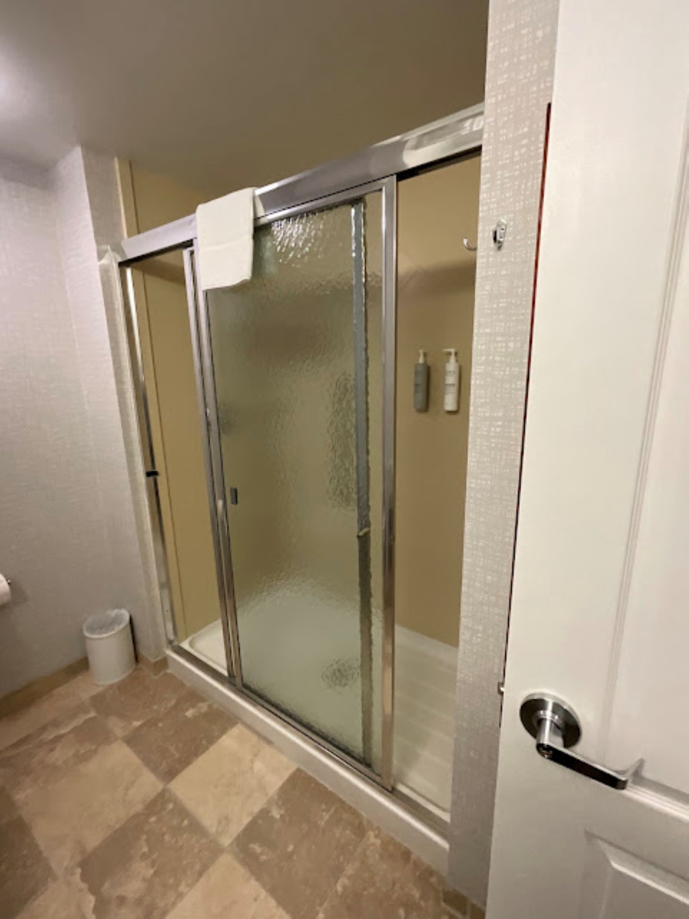 A bathroom with a glass shower enclosure, a white door, and a trash can.