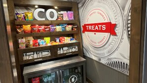 Un présentoir à collations avec une enseigne « CO » et une autre « TREATS », proposant divers en-cas emballés et une section réfrigérée.