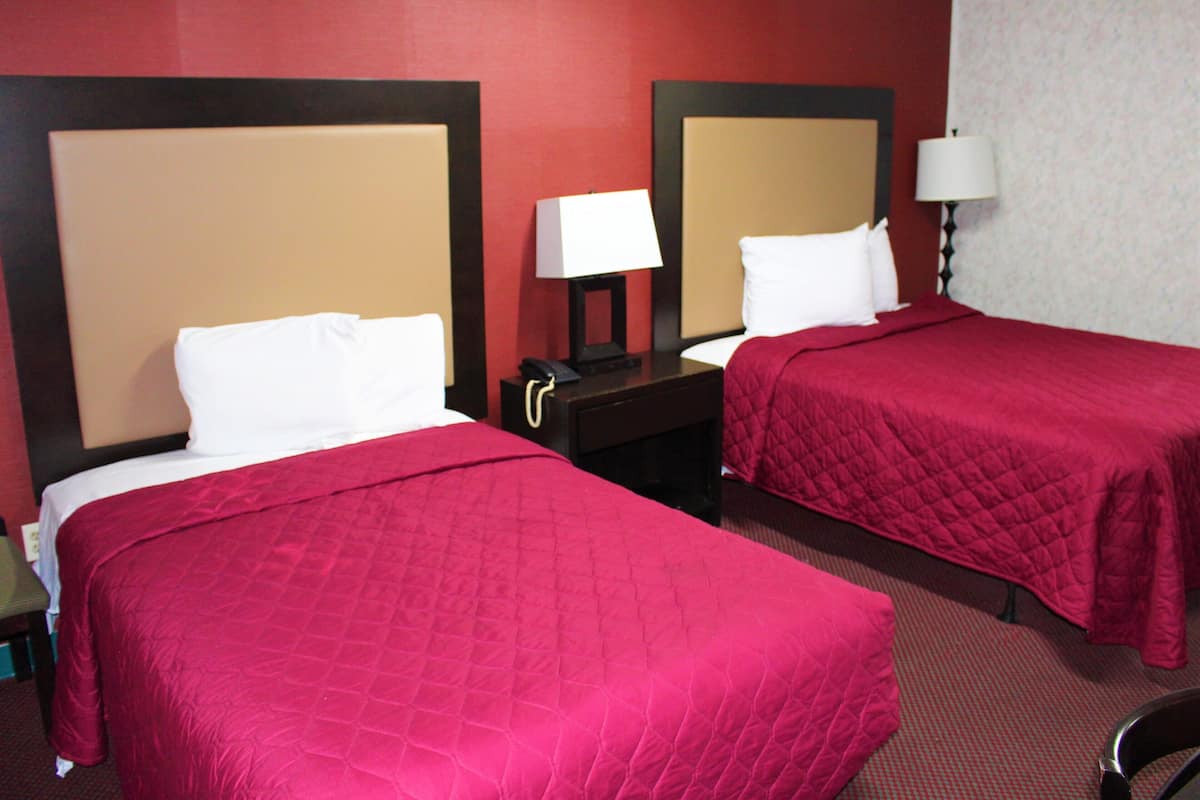 A hotel room with two beds, each with a red bedspread and white pillows. There is a nightstand with a lamp and a phone between the beds.